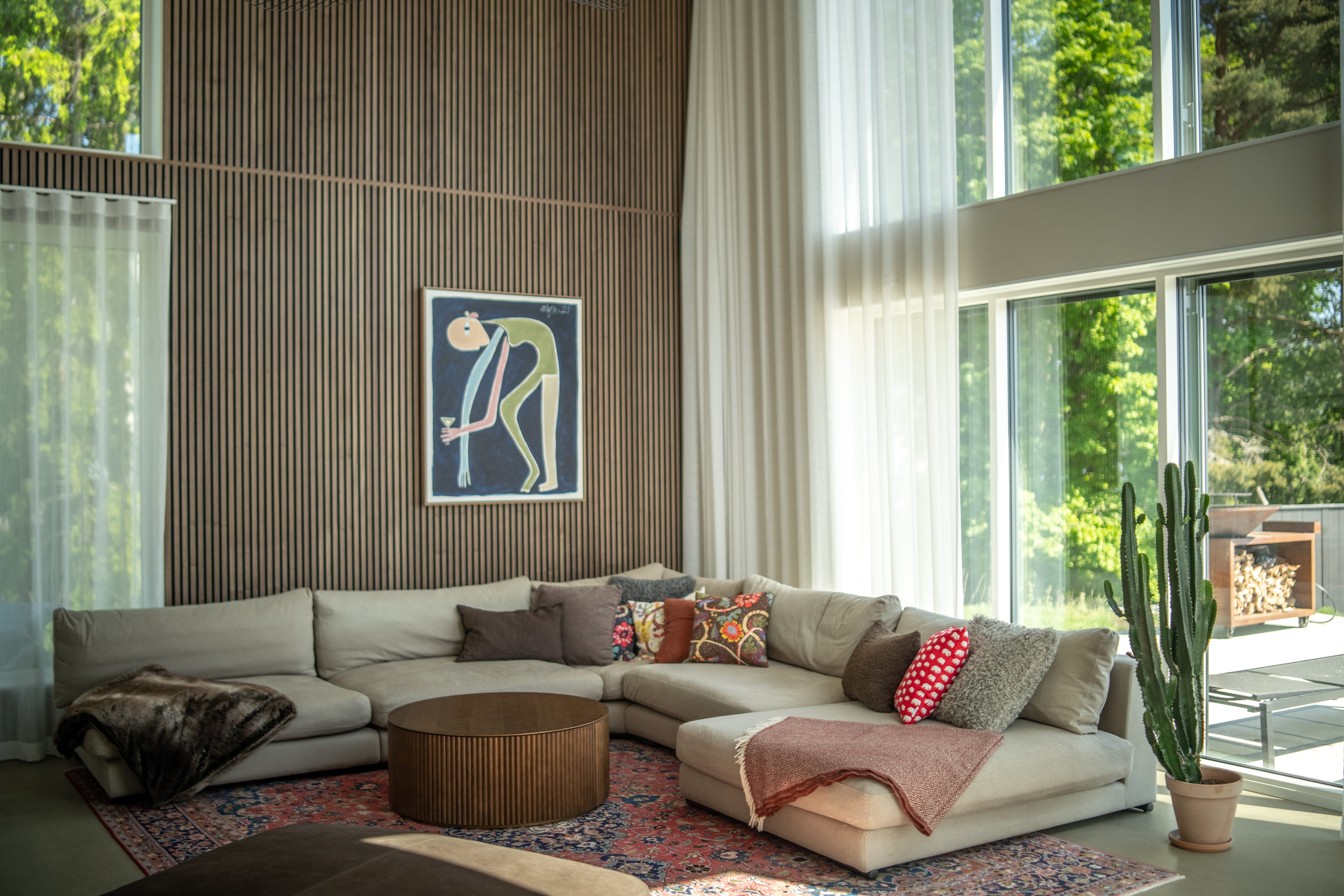 Moderne woonkamer met een grote beige sofa, kleurrijke kussens, ronde salontafel, cactus en ramen van vloer tot plafond met transparante gordijnen en Scandinavische akoestische panelen.
