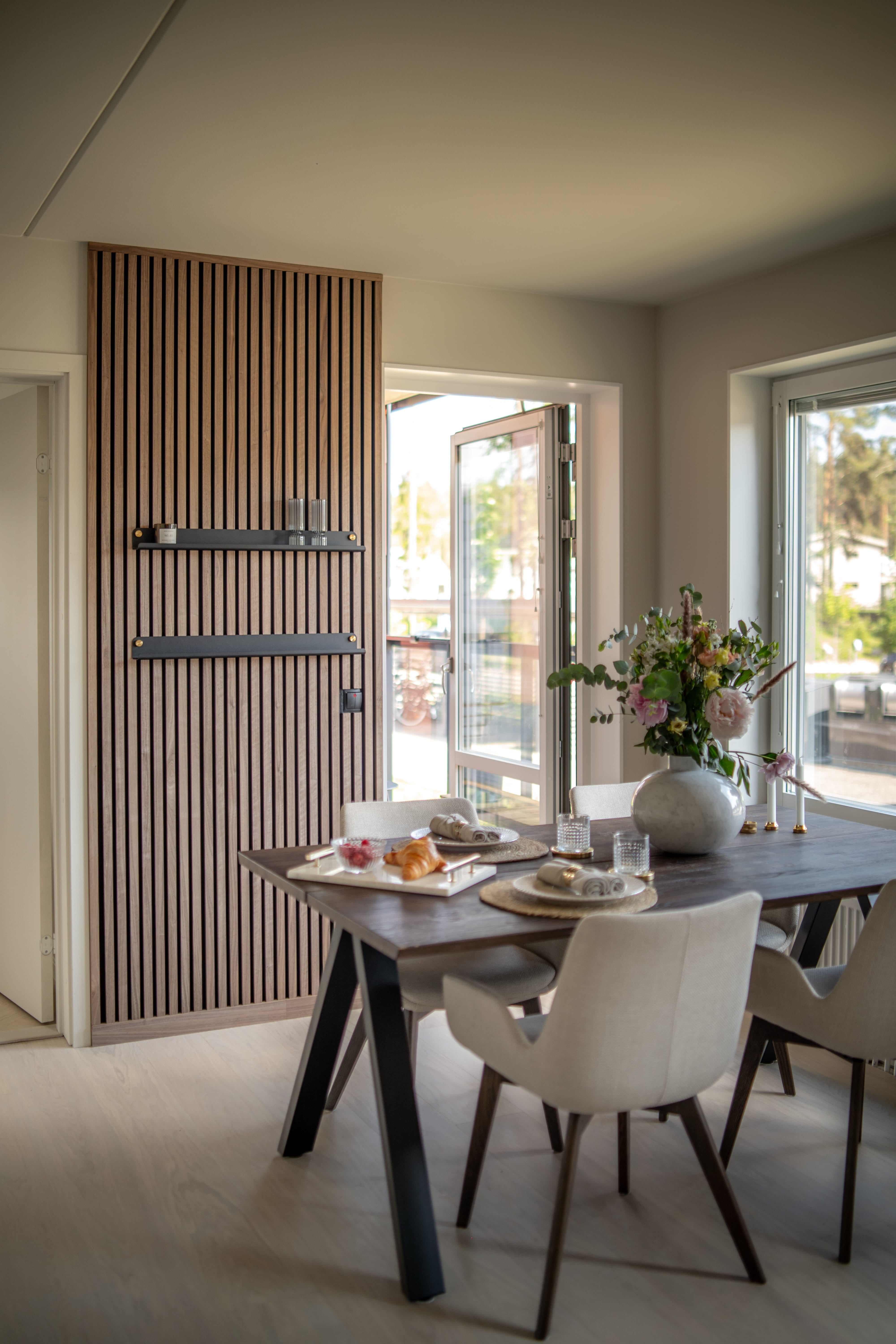 Moderne eetkamer met Akupanel akoestische houten wand met textuur, donkere ontbijttafel, minimalistische beige stoelen en een vaas met bloemen bij grote open ramen.