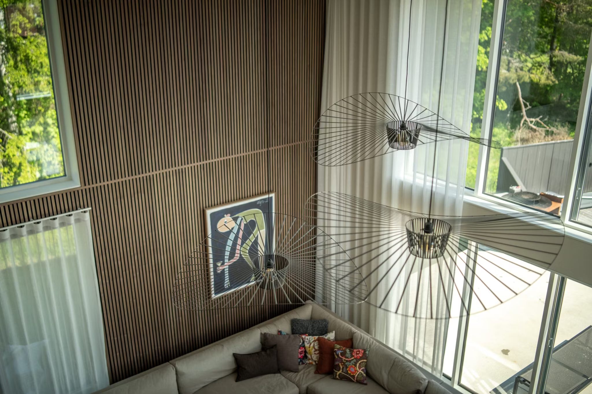 Moderne woonkamer met Scandinavisch houten Akupanel muren, grote ramen, abstracte akoestische paneelkunst, een sofa en minimalistische draadframe verlichtingsarmaturen, van bovenaf gezien.