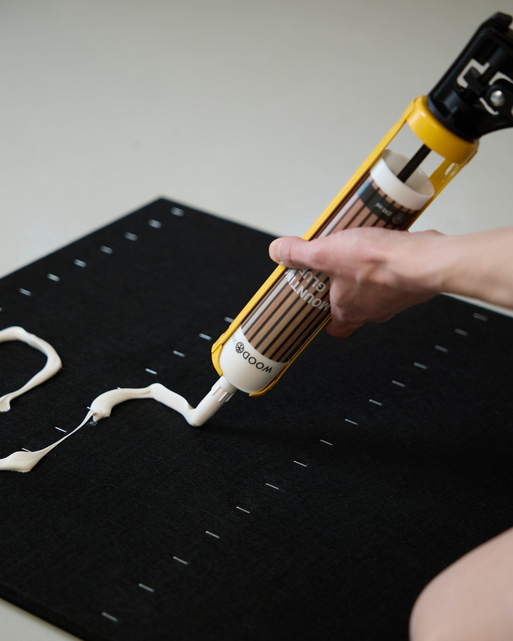 Een hand brengt met het WoodUpp voegkitpistool (290 ml) witte montagelijm in een zigzagpatroon aan op een black Akoestisch paneel oppervlak.