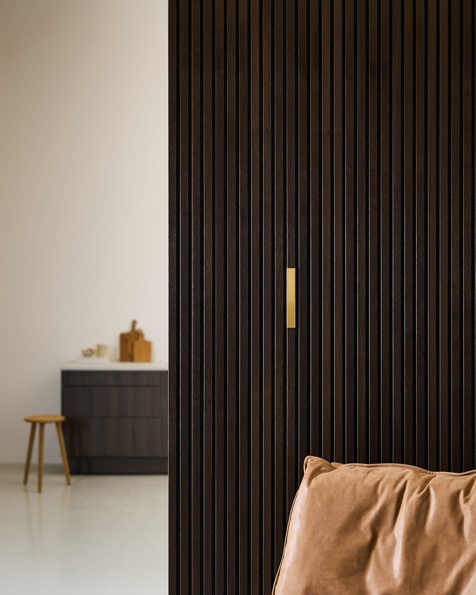 Pièce minimaliste agrémentée dun accessoire WoodUpp Liva | Cover / Laiton, 20 cm, posé sur une cloison Akupanel en bois foncé, dun fauteuil en cuir marron et dun meuble noir.