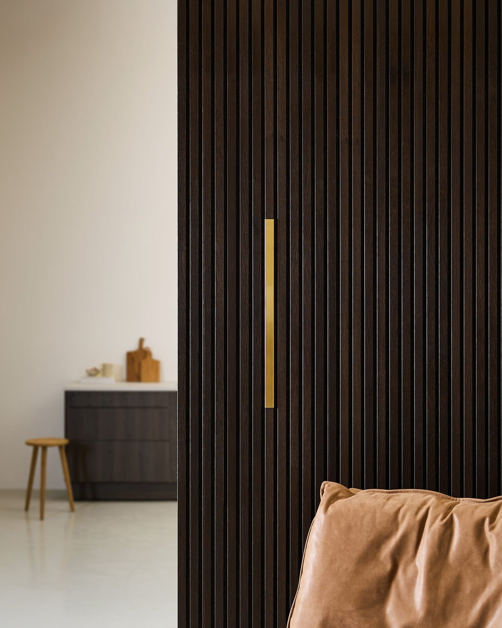 Minimalistische kamer met een WoodUpp Akupanel Liva | Afwerking / Messing, 60 cm, naast een leren stoel, een kruk en een donkere kast in een neutrale omgeving.