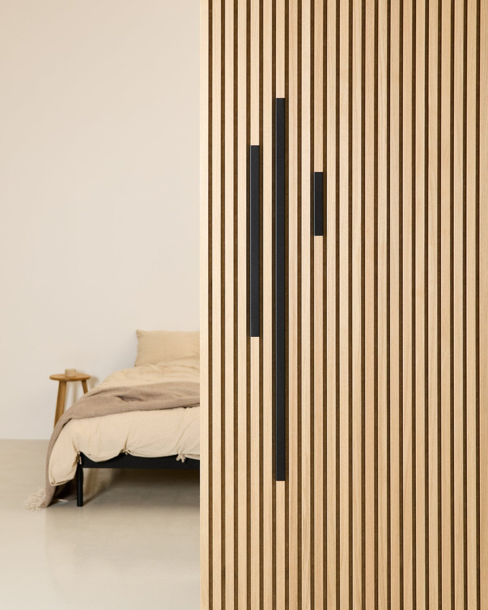 Minimalistische slaapkamer met een beige bed en plaid, deels aan het zicht onttrokken door een 60 cm WoodUpp Akupanel roomdivider in Black aluminium uit de Liva Cover-serie.