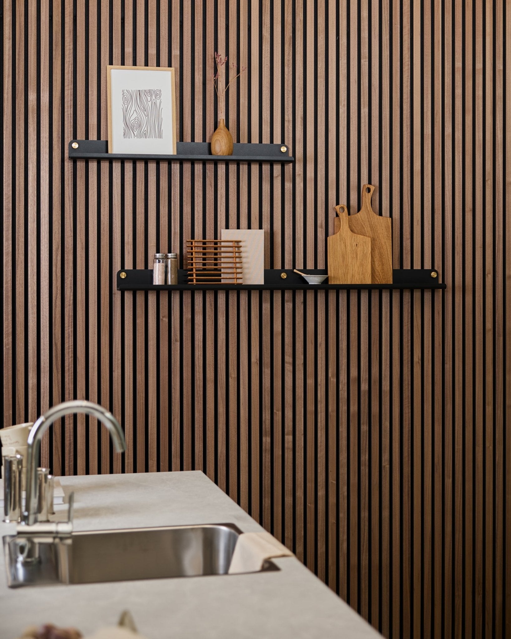 Minimalistische keuken met een vertical Akupanel akoestische houten lattenwand, black planken, snijplanken, small decoratieve items en een modern aanrecht met spoelbak ervoor.