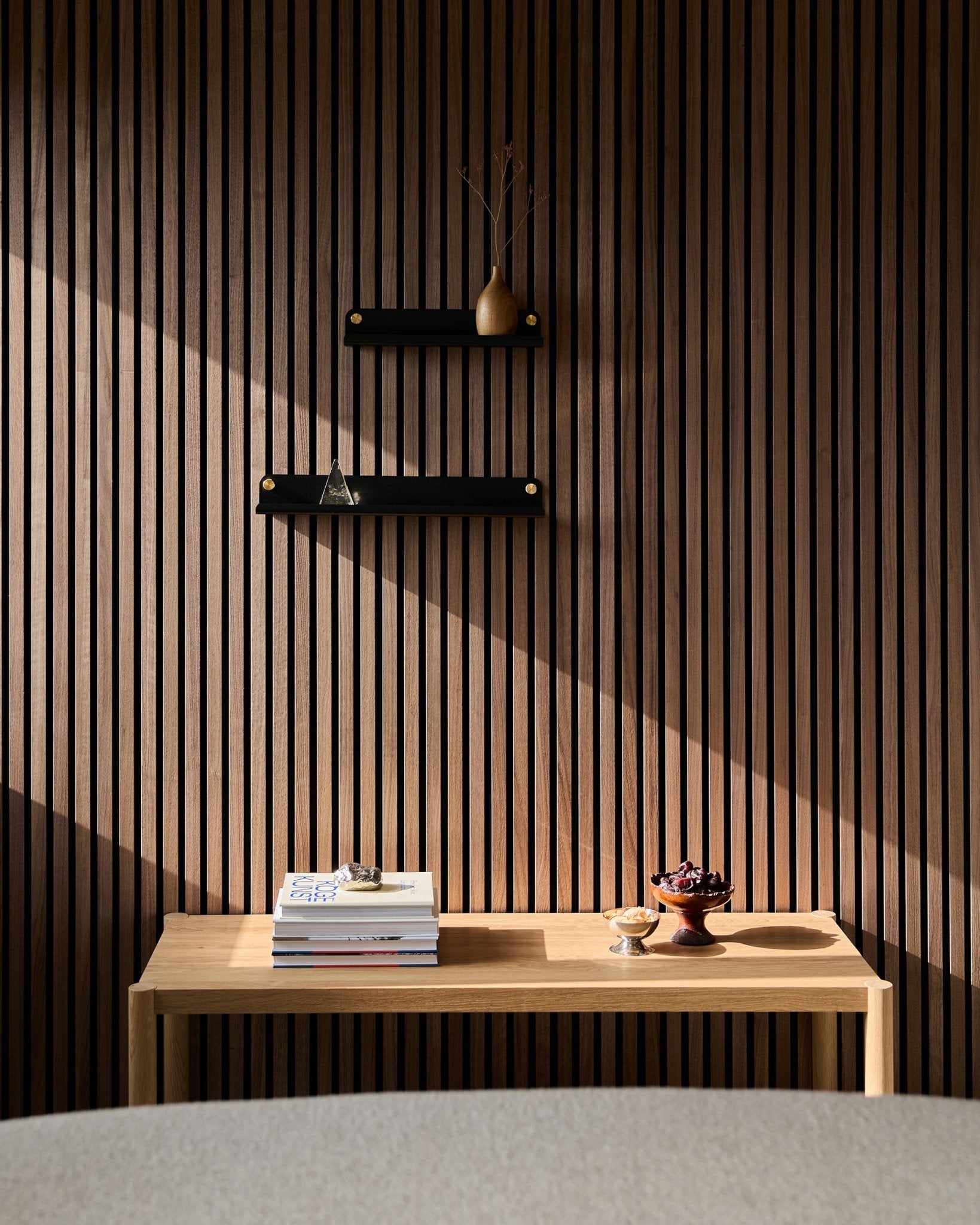 Een minimalistische houten tafel met boeken en kommen staat naast een Nordic lattenwand Akoestisch paneel, met de WoodUpp Norr Narrow Shelf in black (51,9 cm) als subtiel middelpunt; daarboven vallen zonverlichte schaduwen.