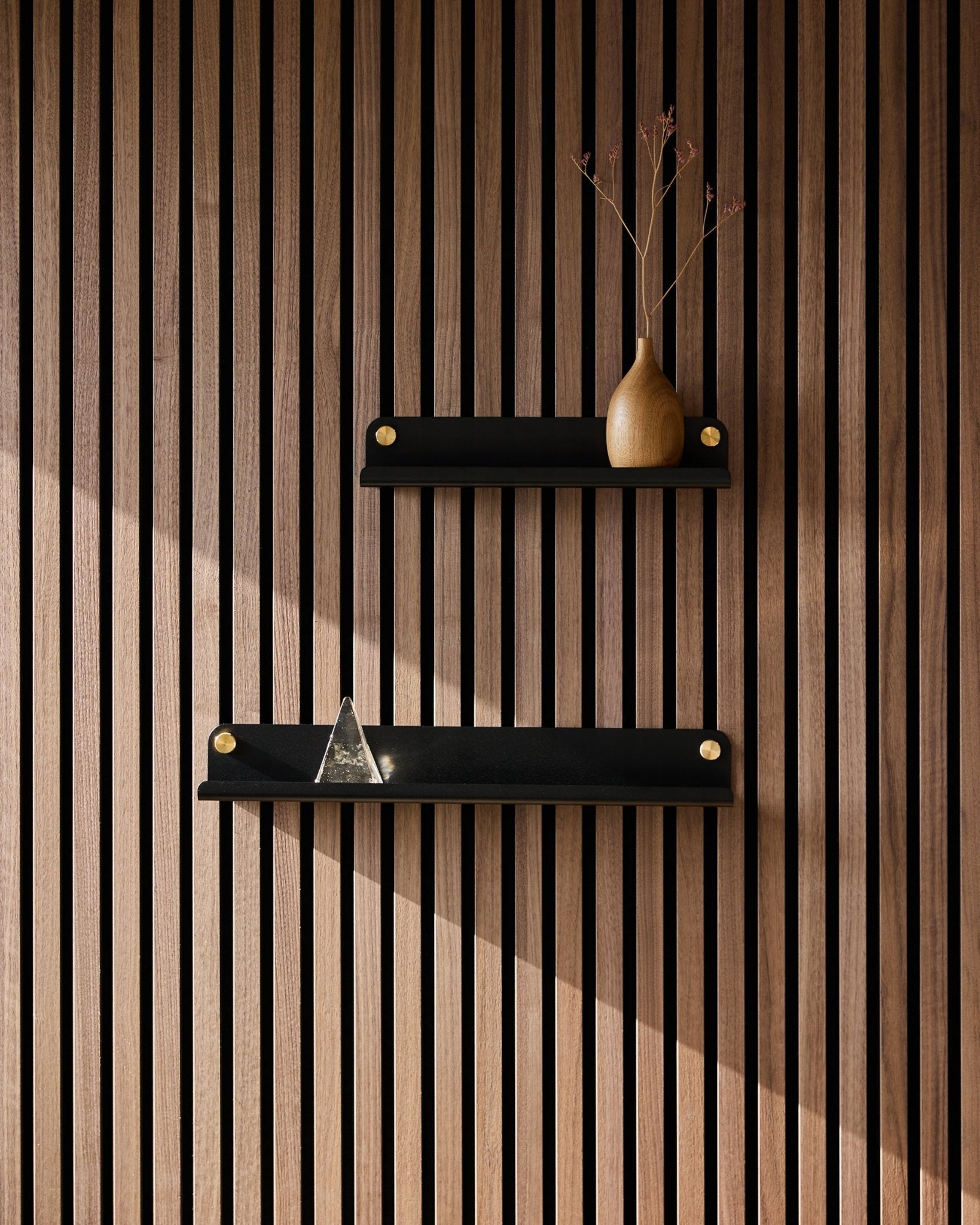 Twee black WoodUpp Norr Narrow shelves (35,9 cm) gemonteerd op een Nordic akoestisch lattenwandpaneel; bovenste plank met een vaas met gedroogde takken, onderste met een glazen piramide.