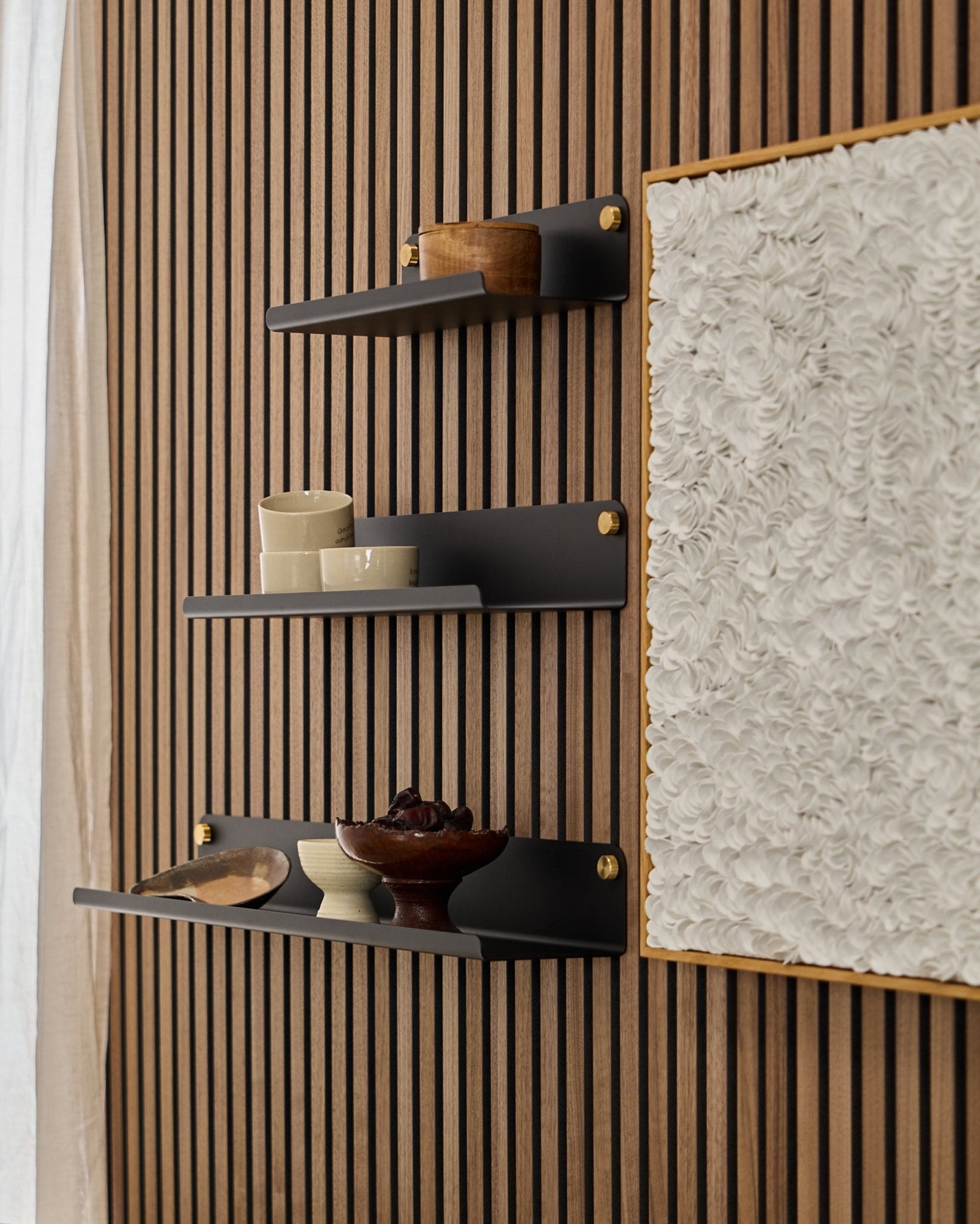 Drie black WoodUpp Norr Wide Shelves (51,9 cm), gemonteerd op een WoodUpp Akupanel akoestisch houten paneel, naast witte kunst met textuur, met kommen en houten containers erop.