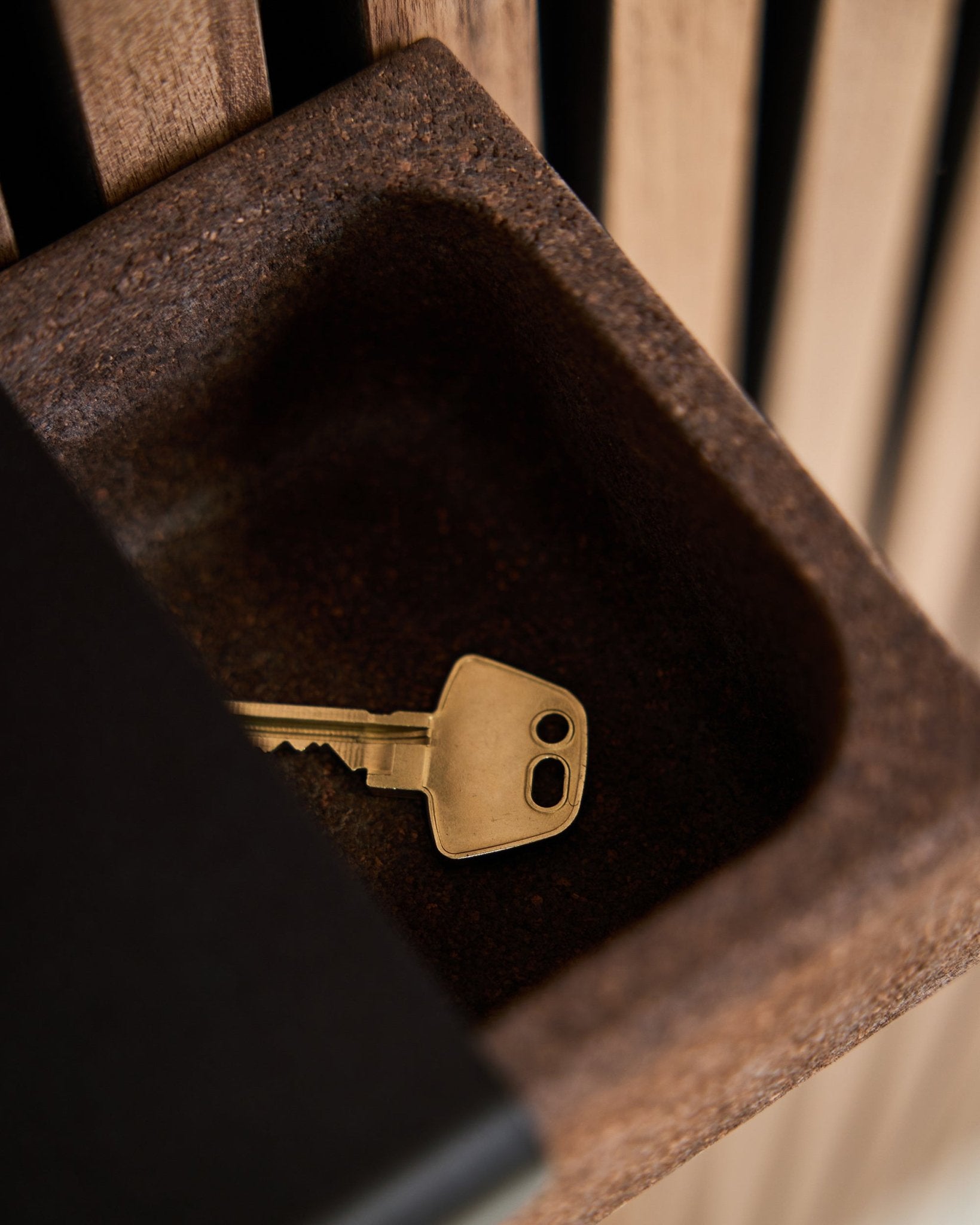 Een brass sleutel ligt in een Norr Key Tray/Smoked Cork, 35,9 cm van WoodUpp, geplaatst op een brown opbergbox voor een achtergrond van akoestische houten panelen in Akupanel.