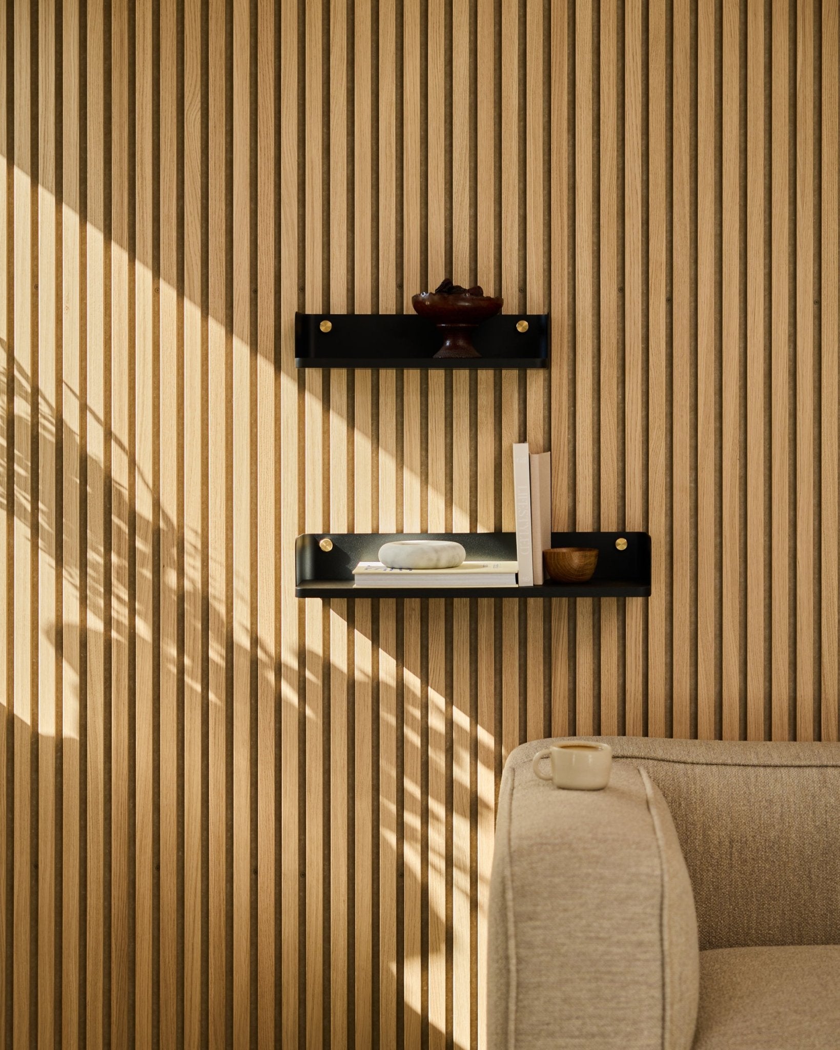 Zonlicht valt op een beige bank en WoodUpps Linn boekenkast in black oak, 55,9 cm, geplaatst tegen een minimalistische Scandinavische lattenwand Akoestisch paneel.