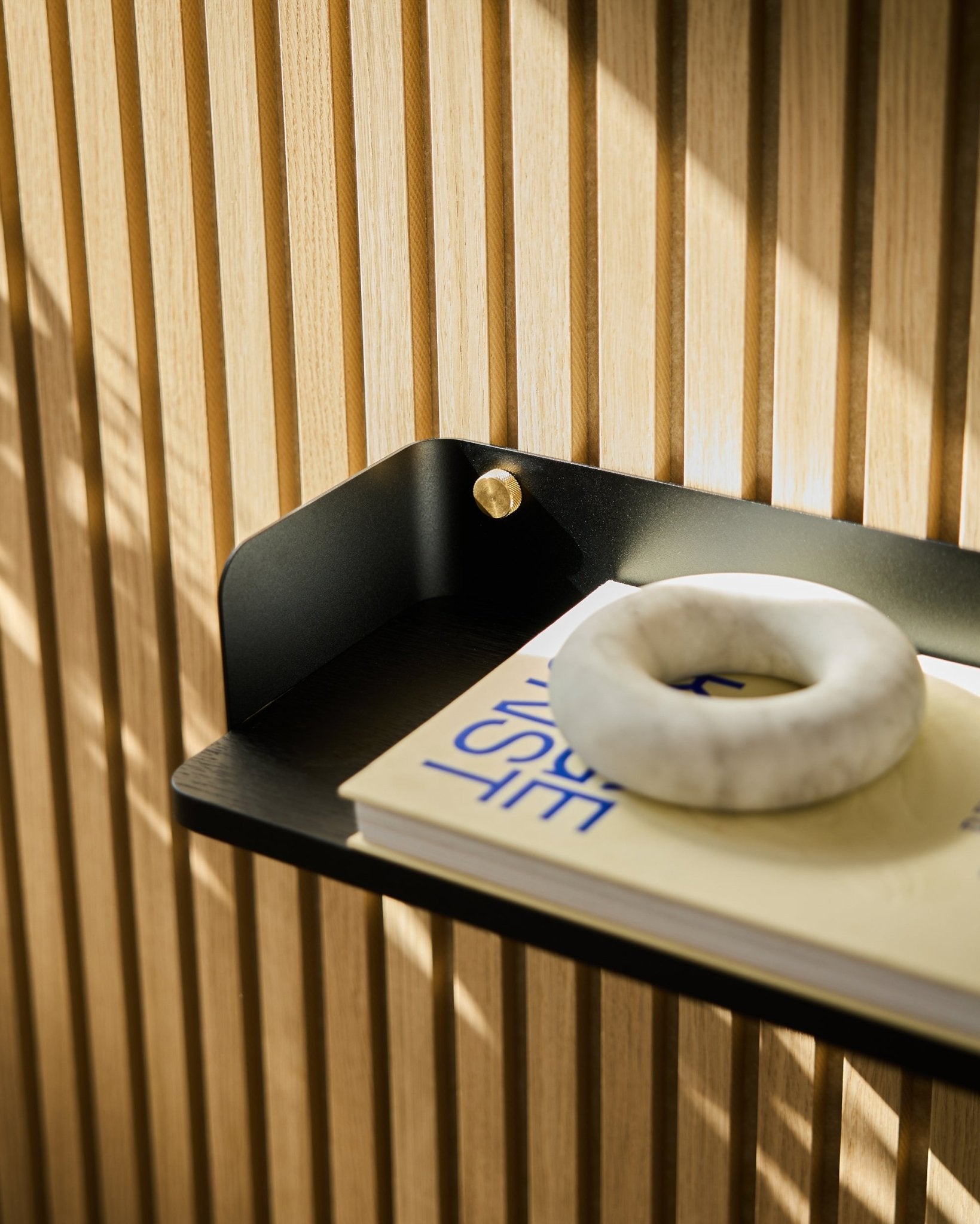 La bibliothèque Black Oak Akupanel de WoodUpp, 55,9 cm, accueille un livre crème avec texte bleu et une pierre ronde grise devant des lattes acoustiques en bois vertical.