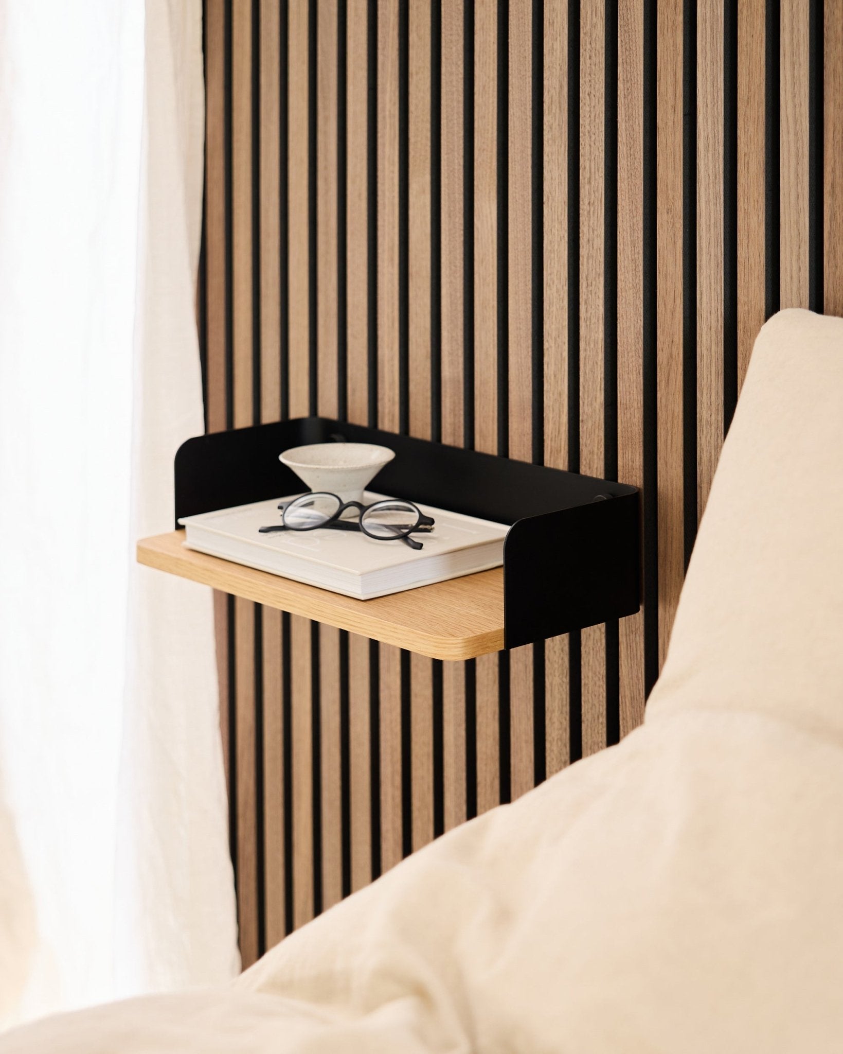 De Linn boekenplank van WoodUpp in natural oak (39,9 cm) met een boek, bril en glas naast een bed tegen een wand met Akupanel akoestische houten panelen.