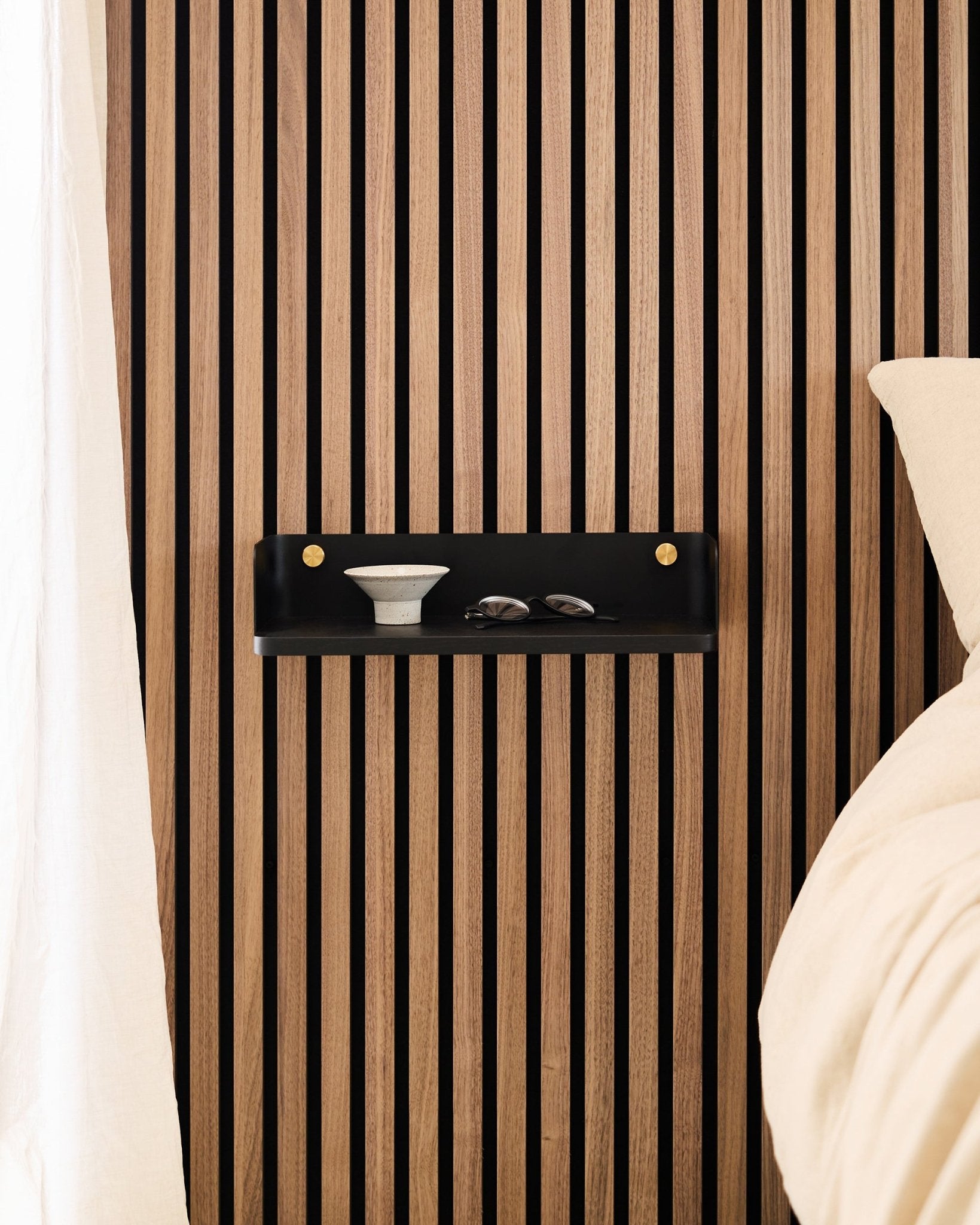 De Linn Bookshelf in Black Oak van WoodUpp is gemonteerd op een Scandinavisch akoestisch lattenwandpaneel en draagt een kom en glazen naast een bed met beddengoed in beige.