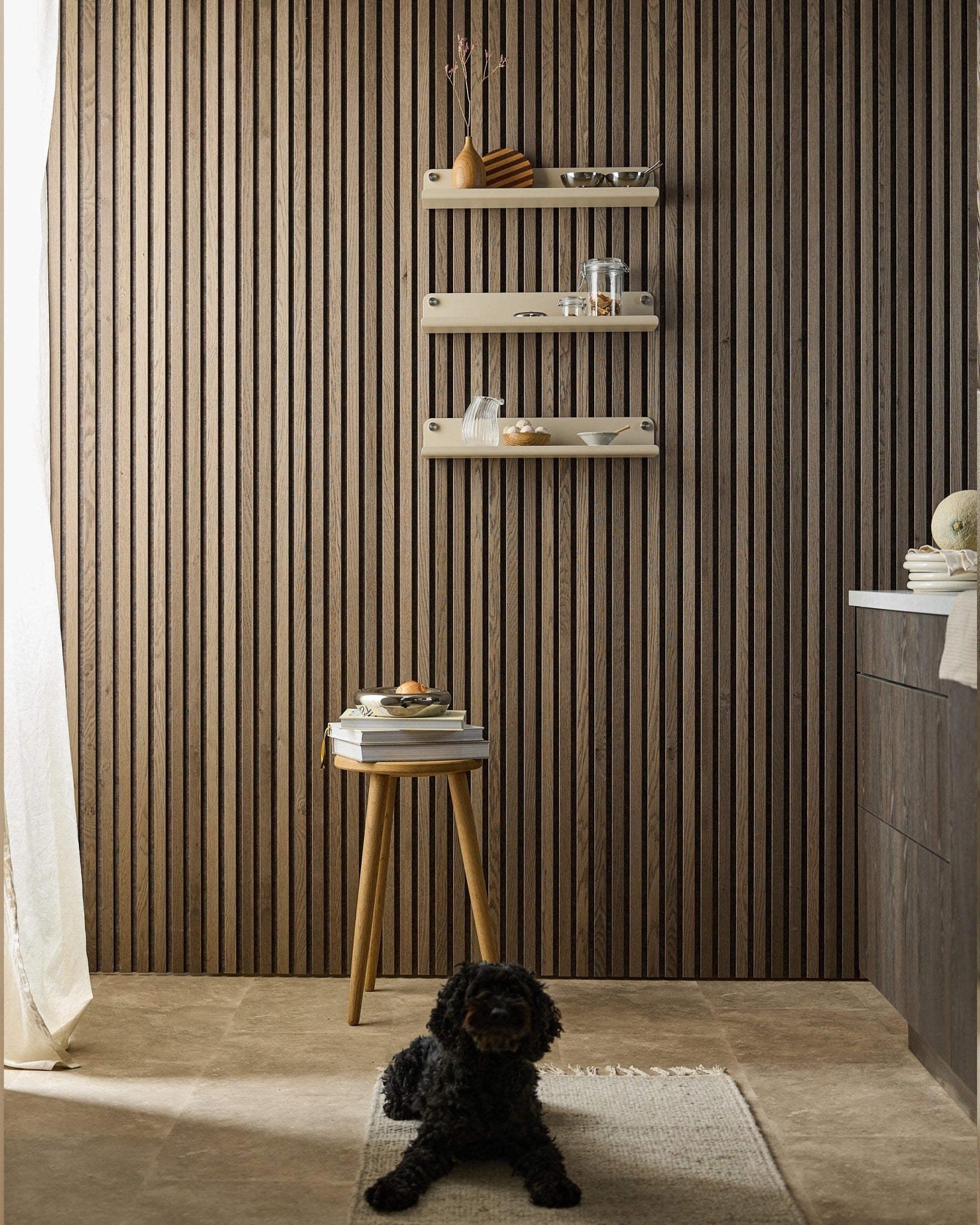 Een black hond ligt op een vloerkleed in een moderne ruimte met een Akupanel Akoestisch paneel wand, een beige WoodUpp Norr brede plank (51,9 cm), een kruk met boeken en zacht licht.