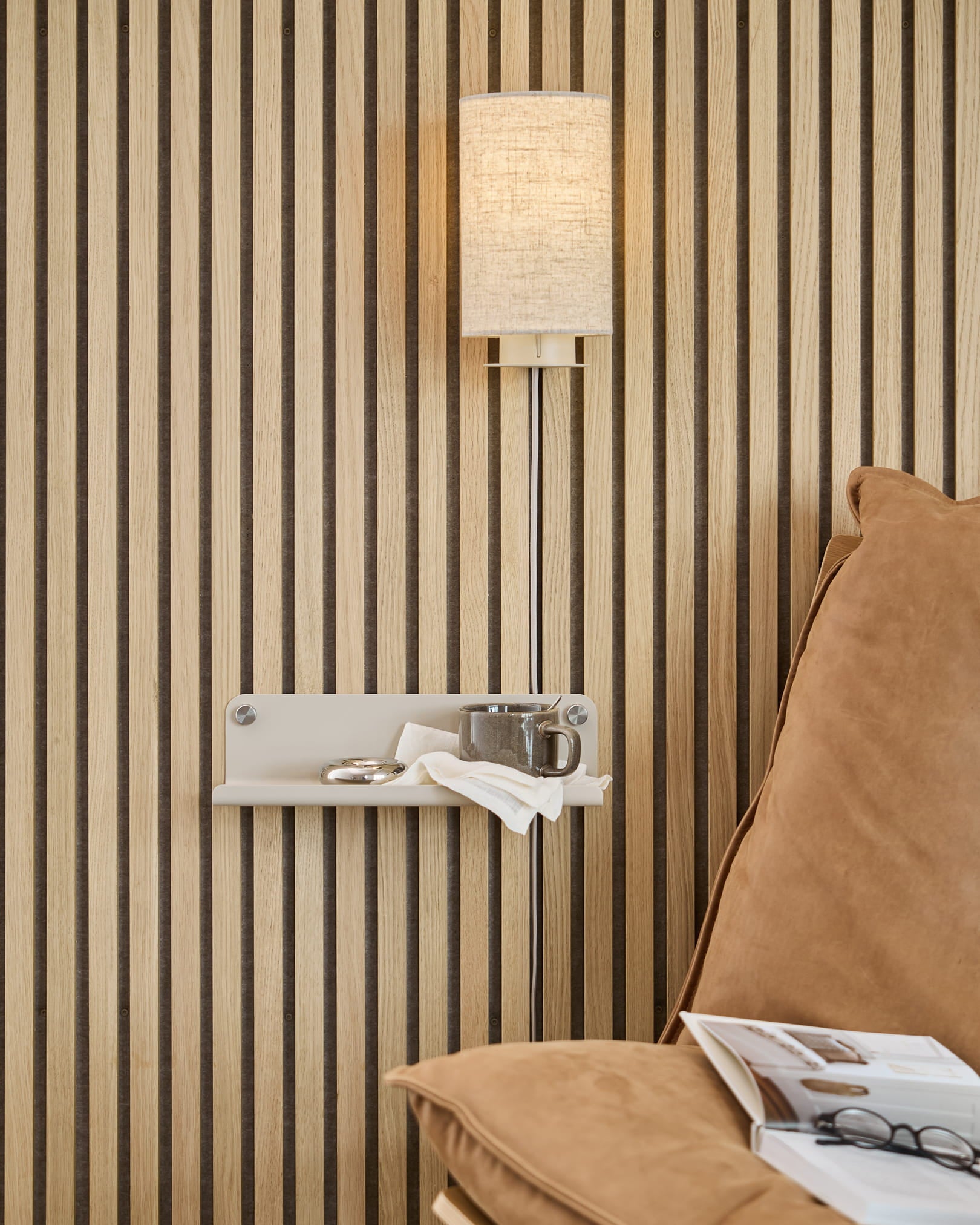 Verticale Akupanel-wand met akoestische houten lamellen, beige wandlamp, klein plankje met een kopje en een servet, bruinbeige bank met een open tijdschrift.