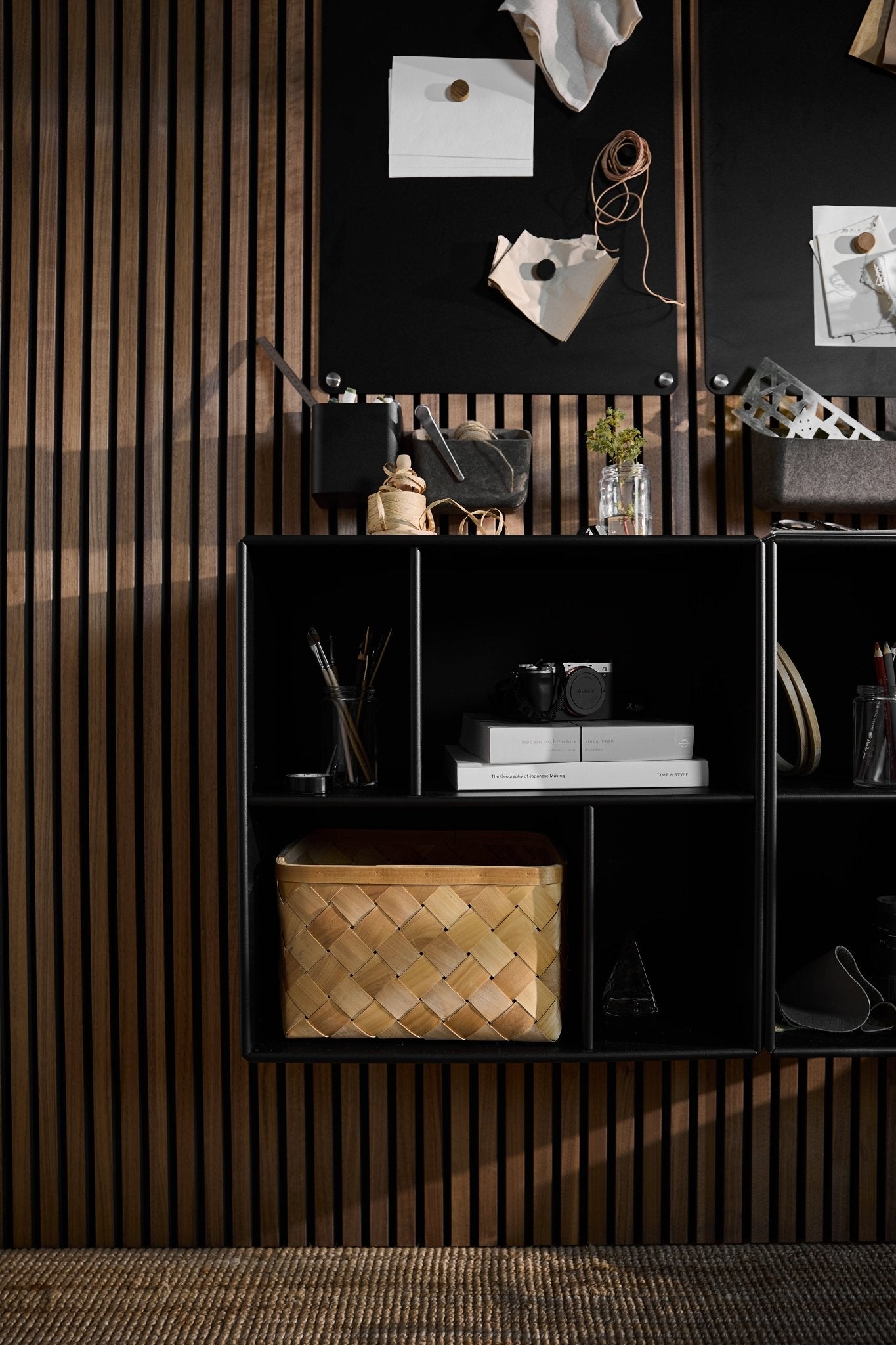 Le meuble ouvert Arka noir de WoodUpp, placé sous un tableau daffichage noir, des livres, un appareil photo, des objets décoratifs et un panier tressé, le tout devant un panneau mural acoustique nordique à lattes.