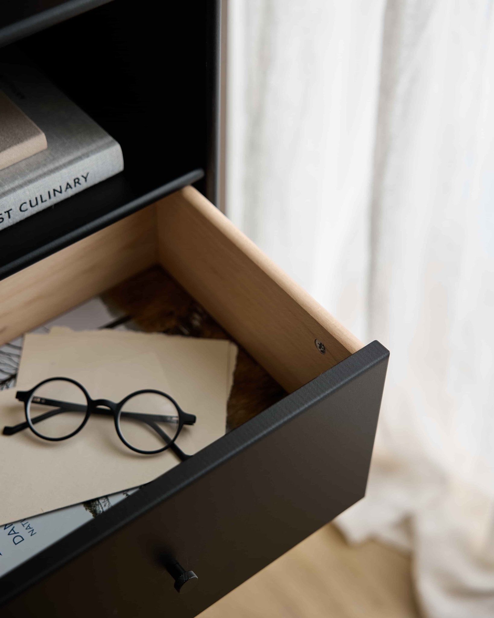 Des lunettes rondes noires et des papiers dans un tiroir ouvert du WoodUpp Arka | Bibliothèque à tiroirs / Noir, sous des livres, avec des rideaux transparents diffusant doucement la lumière ambiante.