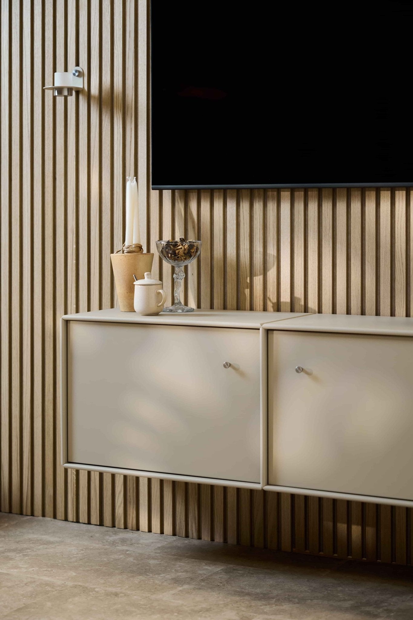 Minimalistische woonkamer met een WoodUpp Arka tv-meubel in beige, een Akupanel akoestische houten wandbekleding, een aan de muur bevestigde tv, kaarsen, een glazen schaal, een witte pot en netjes weggewerkte kabels.