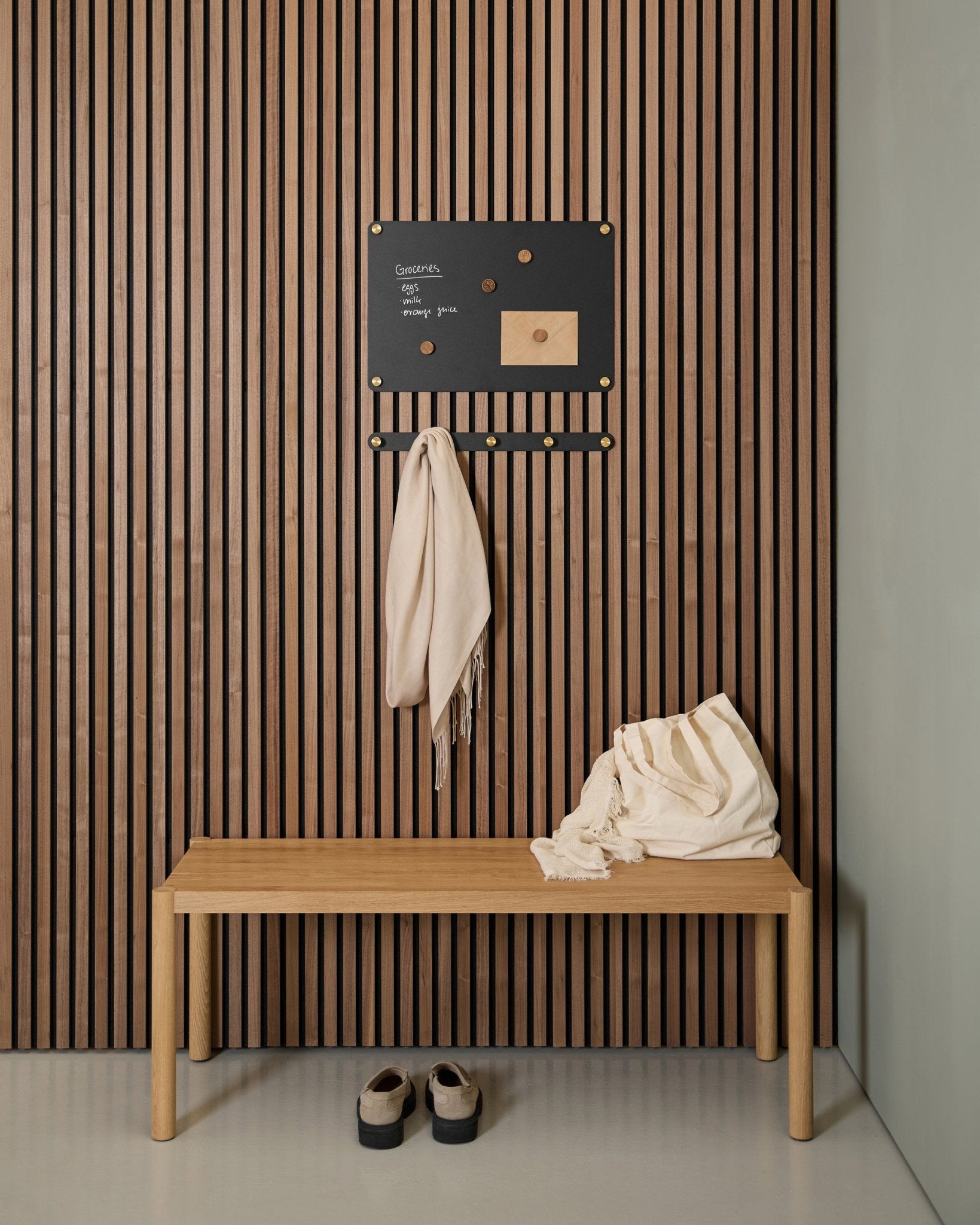 Entrée minimaliste avec un banc en bois, deux écharpes, des sandales et un tableau magnétique WoodUpp Norr small sur un panneau acoustique mural en lattes de bois Akupanel.