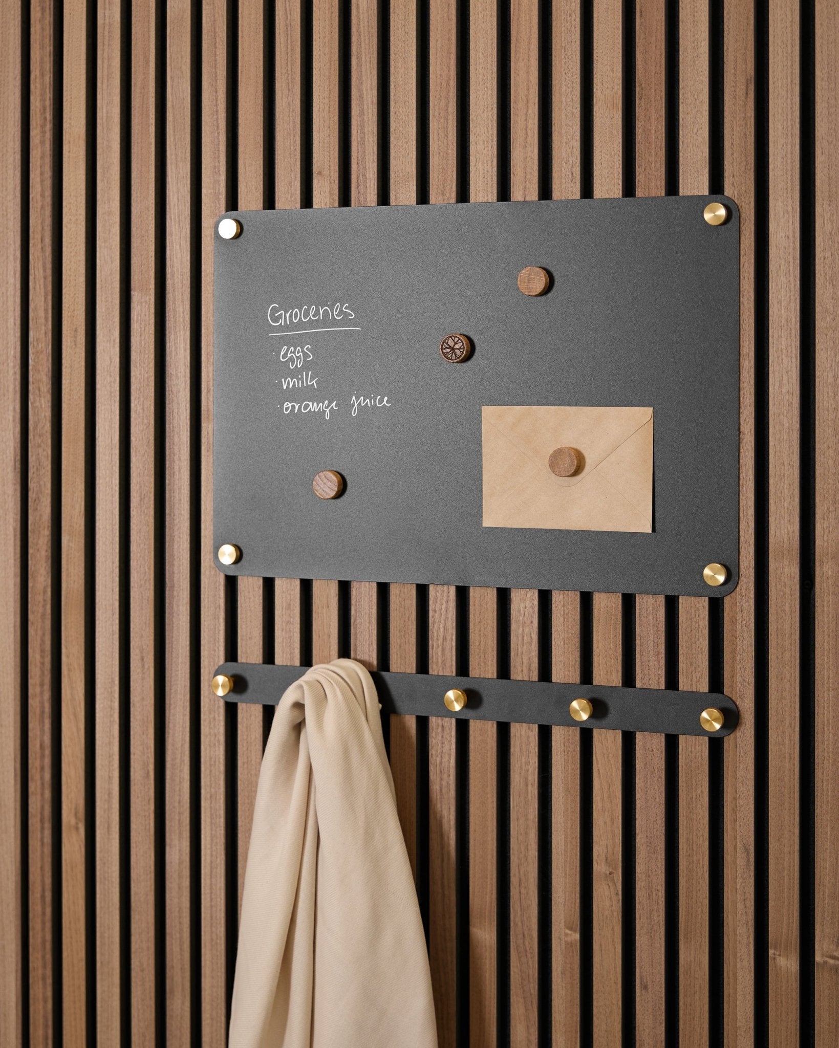 Een small black Norr Magnetic Board van WoodUpp houdt een boodschappenlijst, envelop, magneten en beige doek aan haken vast, gemonteerd op een Akupanel Akoestisch paneel.