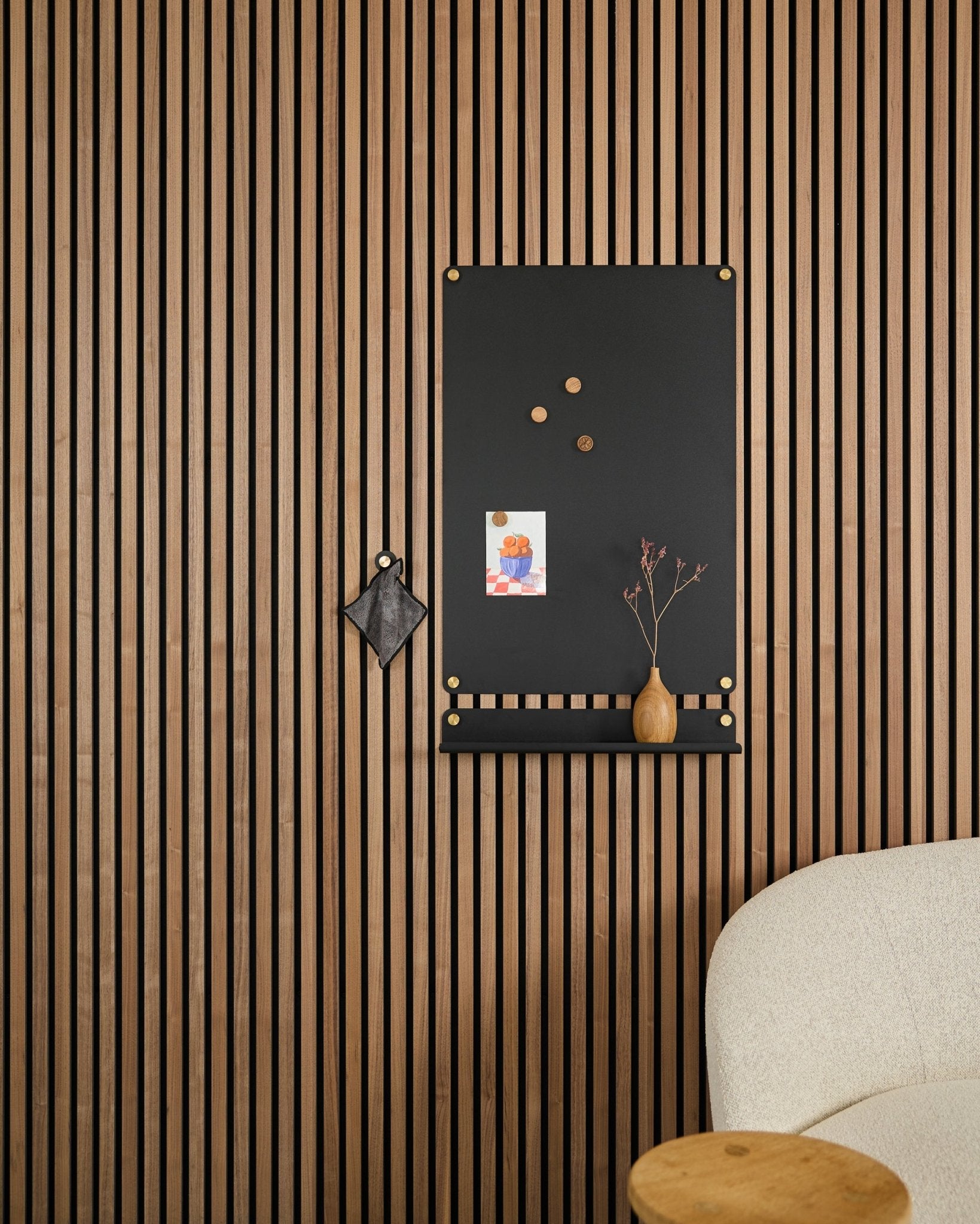 Een large black Norr magnetisch bord van WoodUpp, ontworpen voor akoestische panelen, hangt naast een witte gestoffeerde stoel met notities, een vaas en een klein decoratief object.