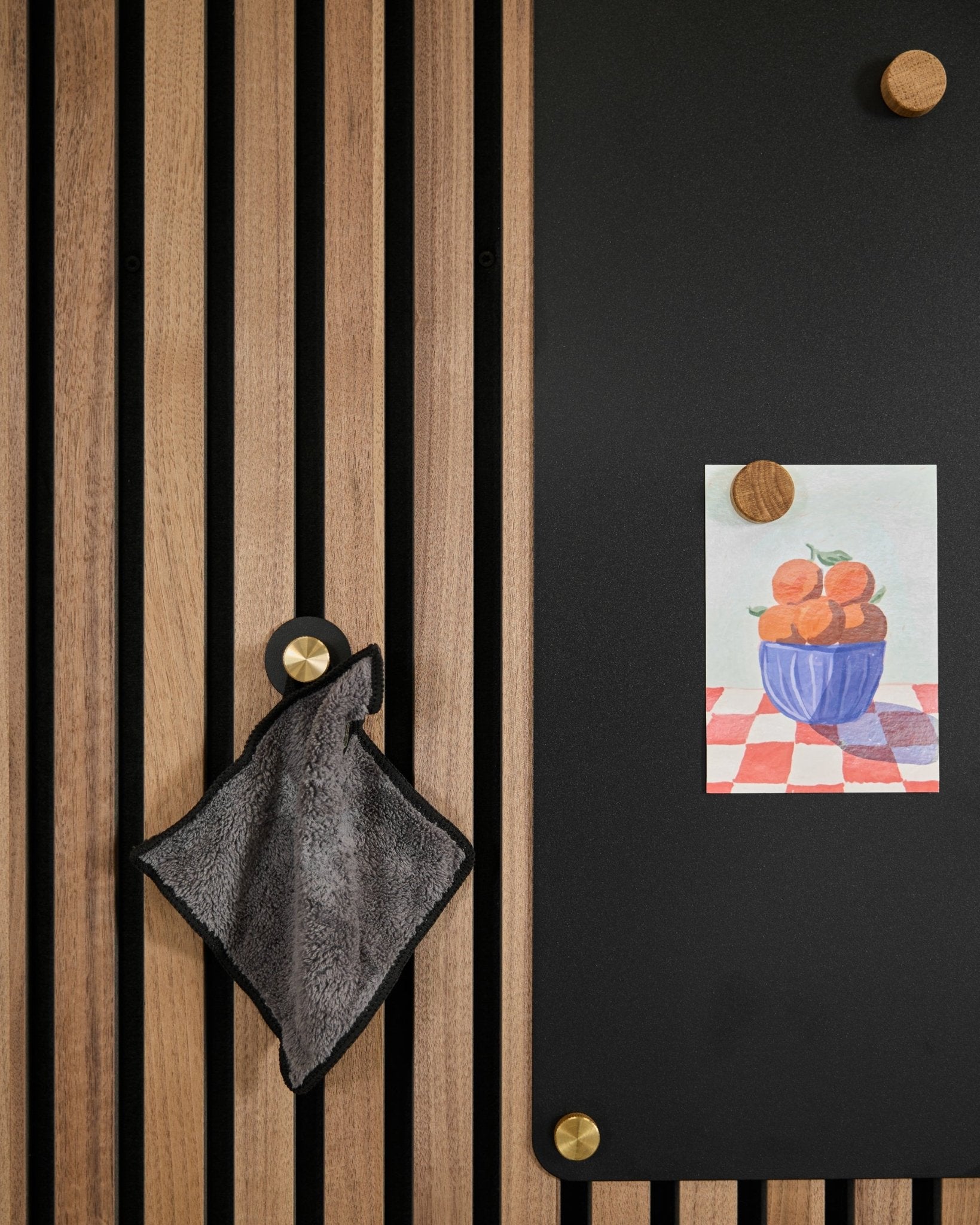 Een small handdoek hangt aan een Nordic akoestisch lattenwandpaneel naast een black bord met een tekening van sinaasappels in een kom, bevestigd met twee gouden magneten.