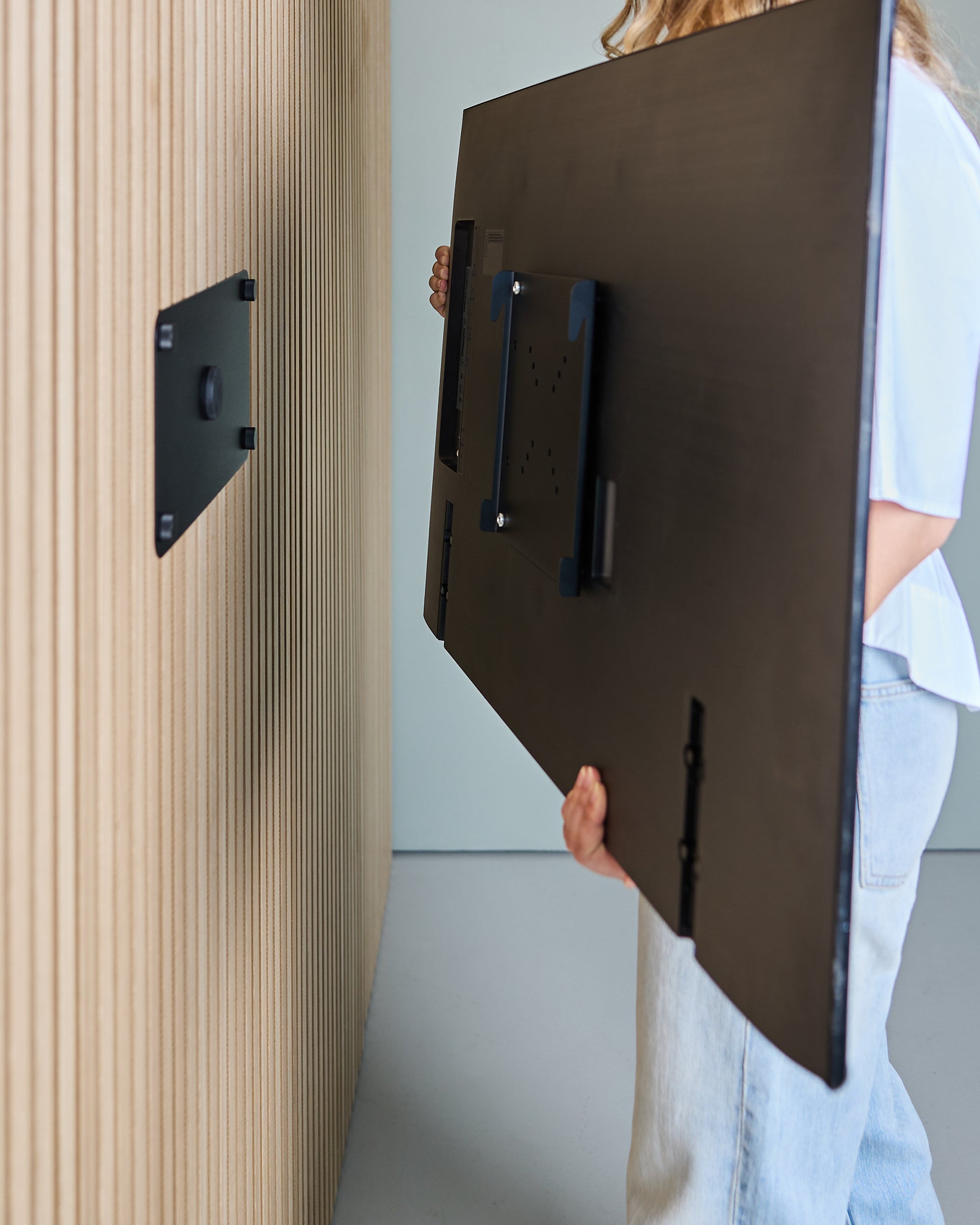 Person holding a flat-screen TV, preparing to mount it on a modern Akupanel acoustic wood slat panel wall.
