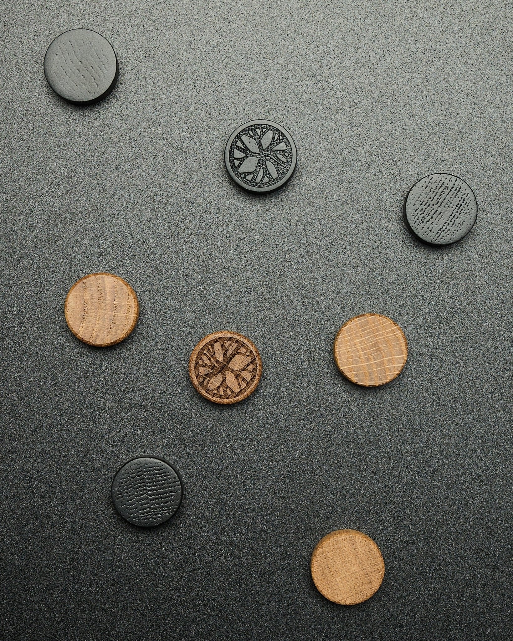 Vier ronde black oak magneten uit de WoodUpp Norr Magnet Set, losjes gerangschikt op een donkere ondergrond met textuur, ontworpen als Akupanel accessoires.