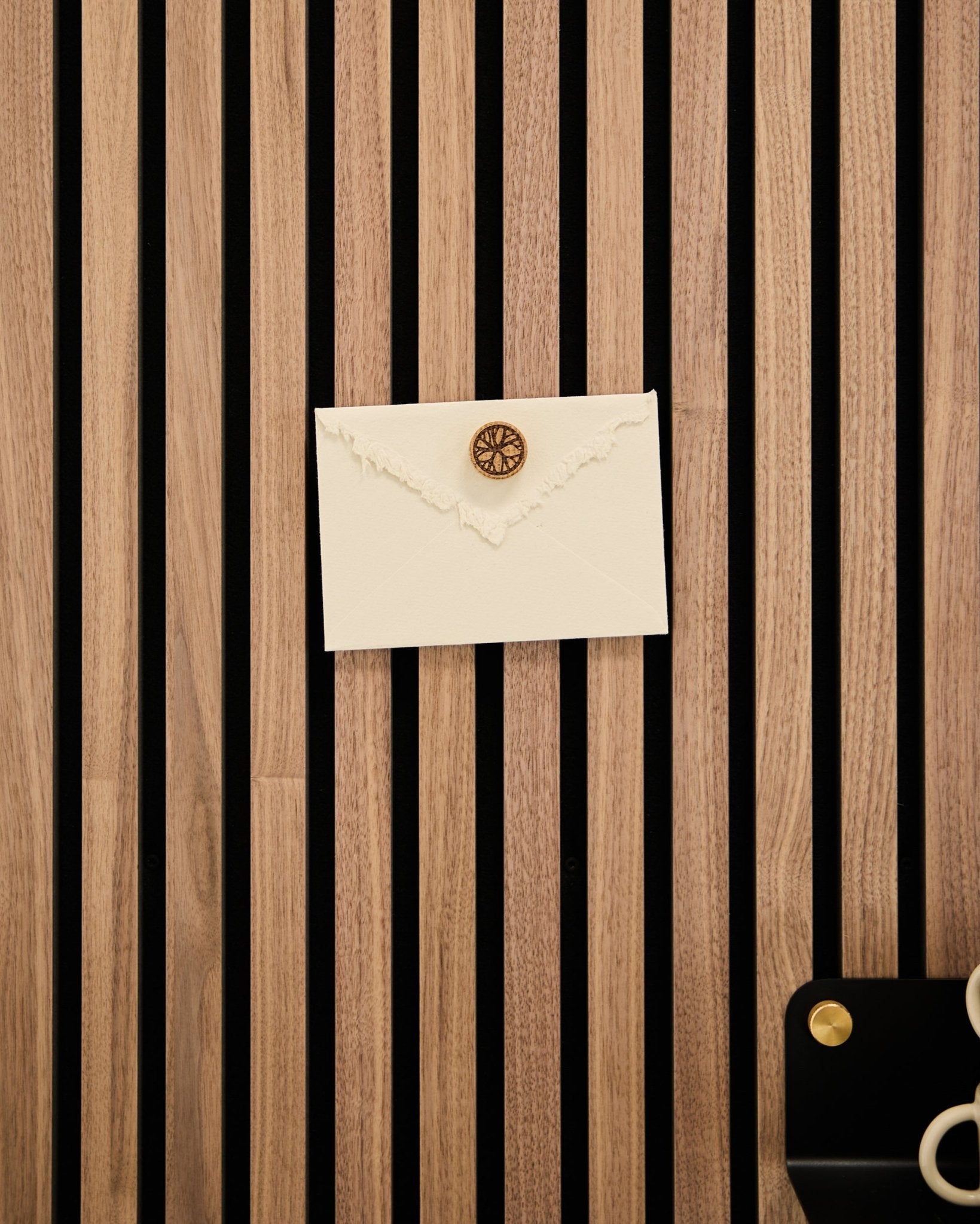Een crèmekleurige envelop met lakzegel is met de Norr Poster Magnet set in Natural Oak bevestigd aan een WoodUpp eiken akoestisch lattenwandpaneel; schaar en schaal liggen in de buurt.