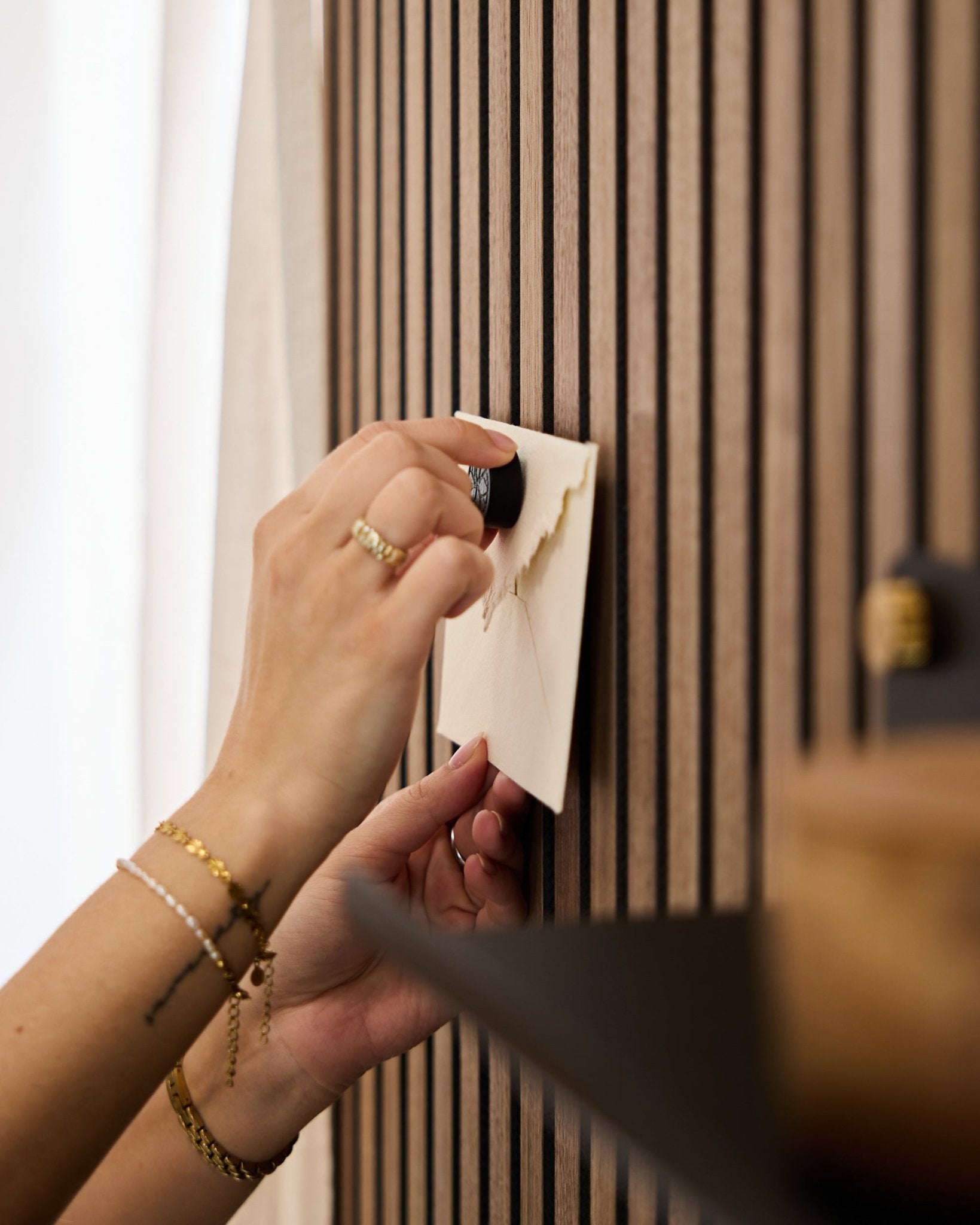 Iemand met gouden armbanden en ringen bevestigt een envelop aan een akoestisch eiken lattenwandpaneel van WoodUpp met de Norr Poster Magnet-set in Black Oak.