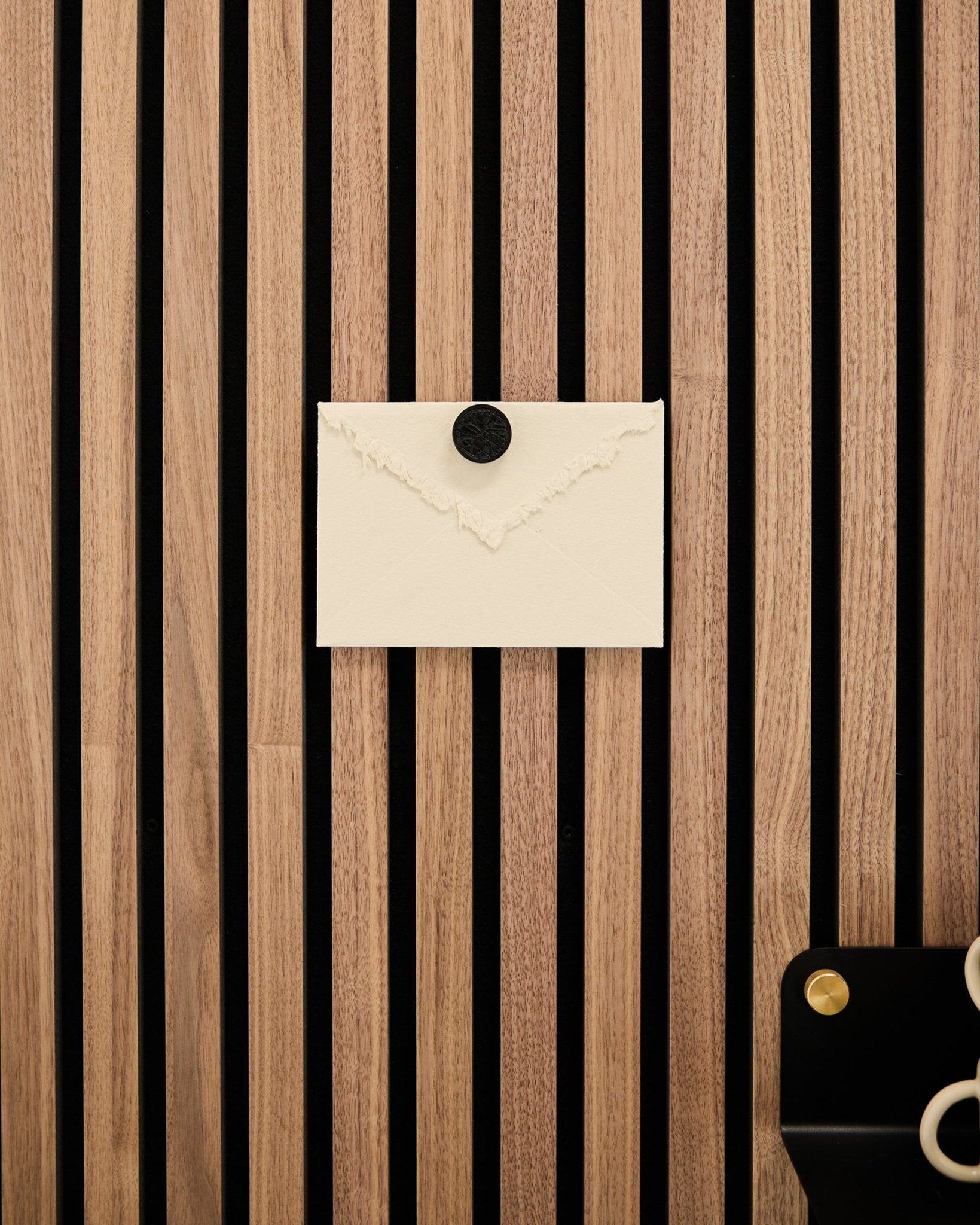 Een crèmekleurige envelop is met de Norr Poster Magnet-set in Black Oak vastgemaakt aan een WoodUpp Akupanel eiken lattenwandpaneel Akoestisch paneel; rechtsonder ligt een schaar.
