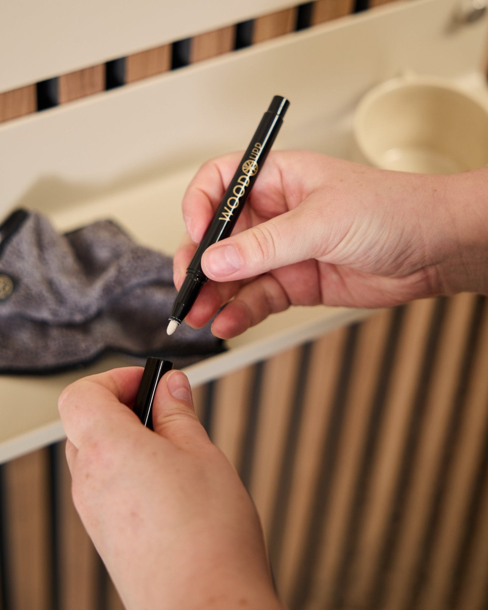 Une personne tient un marqueur WoodUpp Norr | Blackboard set ouvert à côté d’un chiffon en microfibre et d’une tasse beige, des produits conçus pour être utilisés avec des panneaux acoustiques en bois.