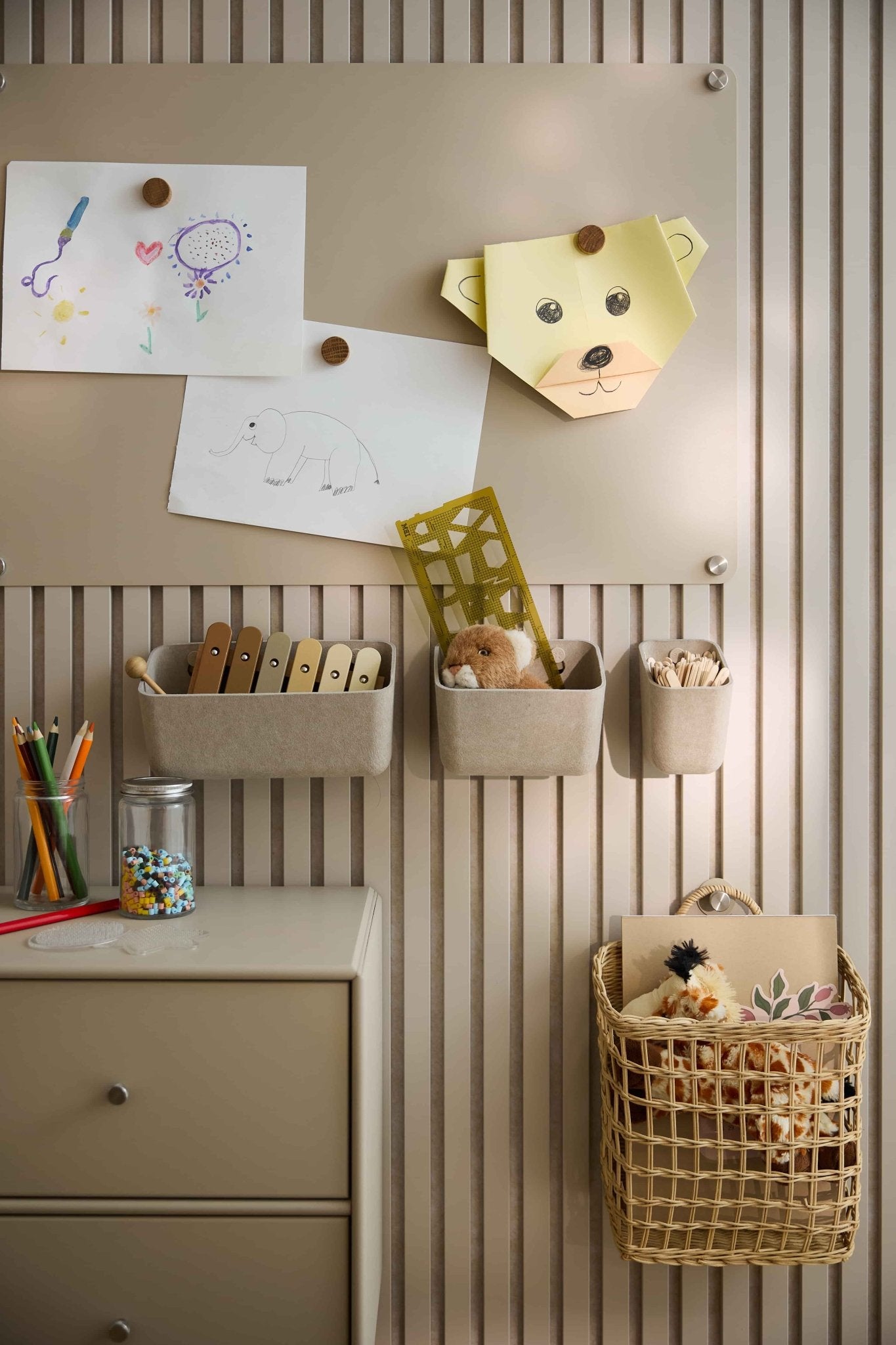 Les dessins et les jouets des enfants sont disposés sur un mur beige recouvert dun panneau acoustique WoodUpp, comprenant des bacs en feutre, des paniers, un tiroir et un panneau perforé.