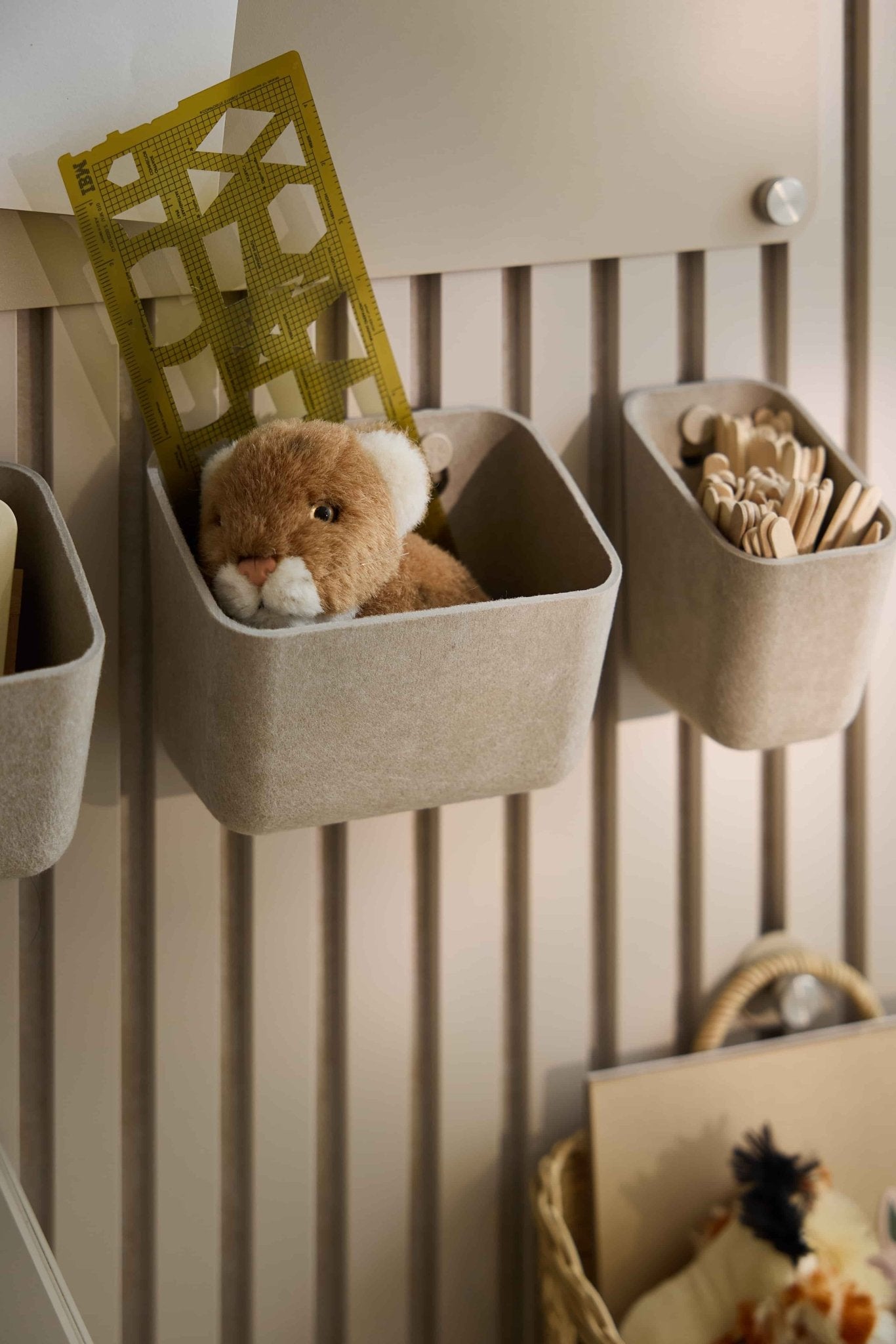 In de beige WoodUpp Luva-viltbakjes worden speelgoed, een gele liniaal en houten stokjes bewaard, zodat alles netjes opgeborgen is op een gestreept akoestisch lamellenpaneel.