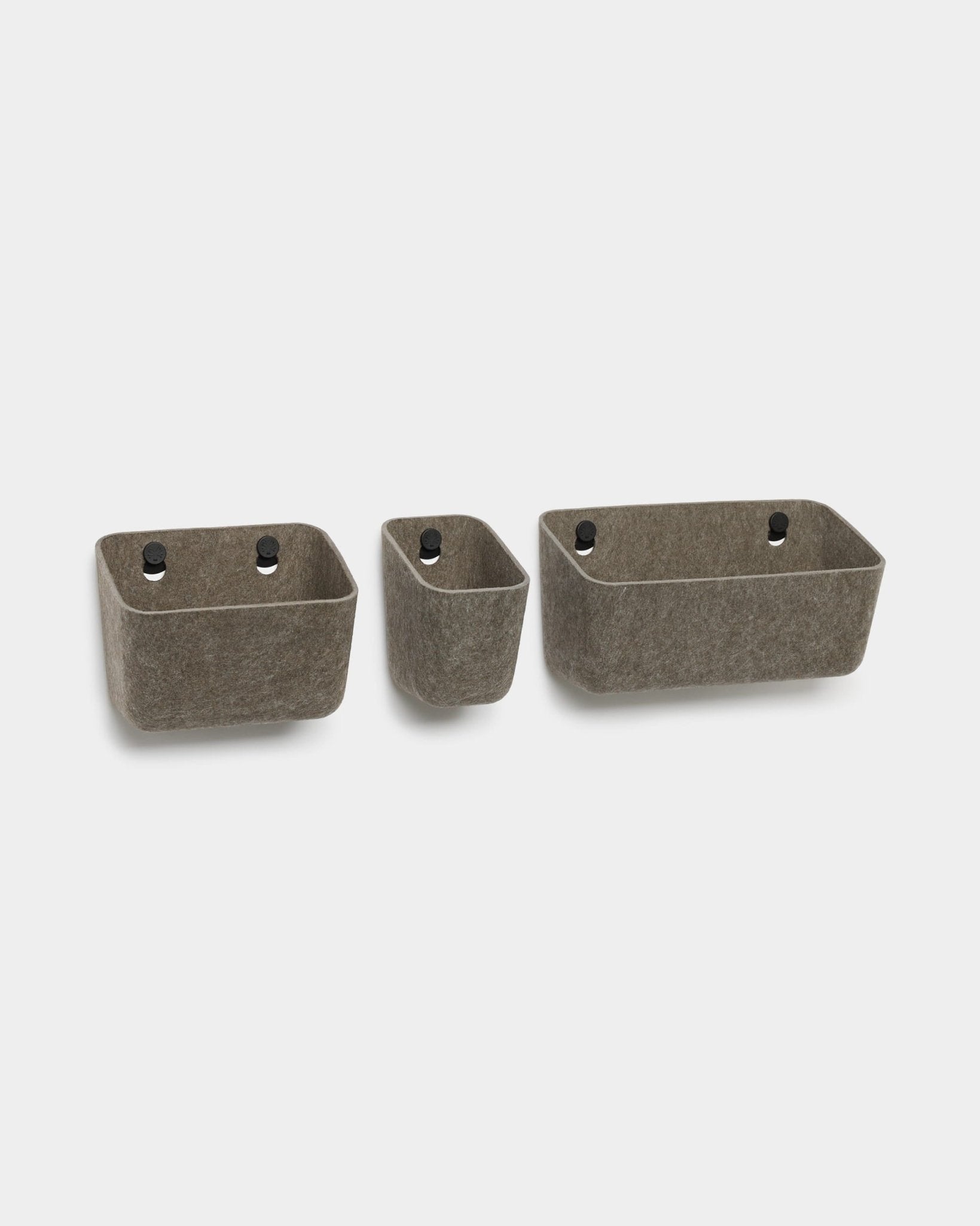 Trois bacs en feutre WoodUpp Luva, de couleur gris foncé chaud, dotés dœillets noirs, conçus pour être fixés au mur et servir de rangements compatibles avec les panneaux acoustiques WoodUpp.