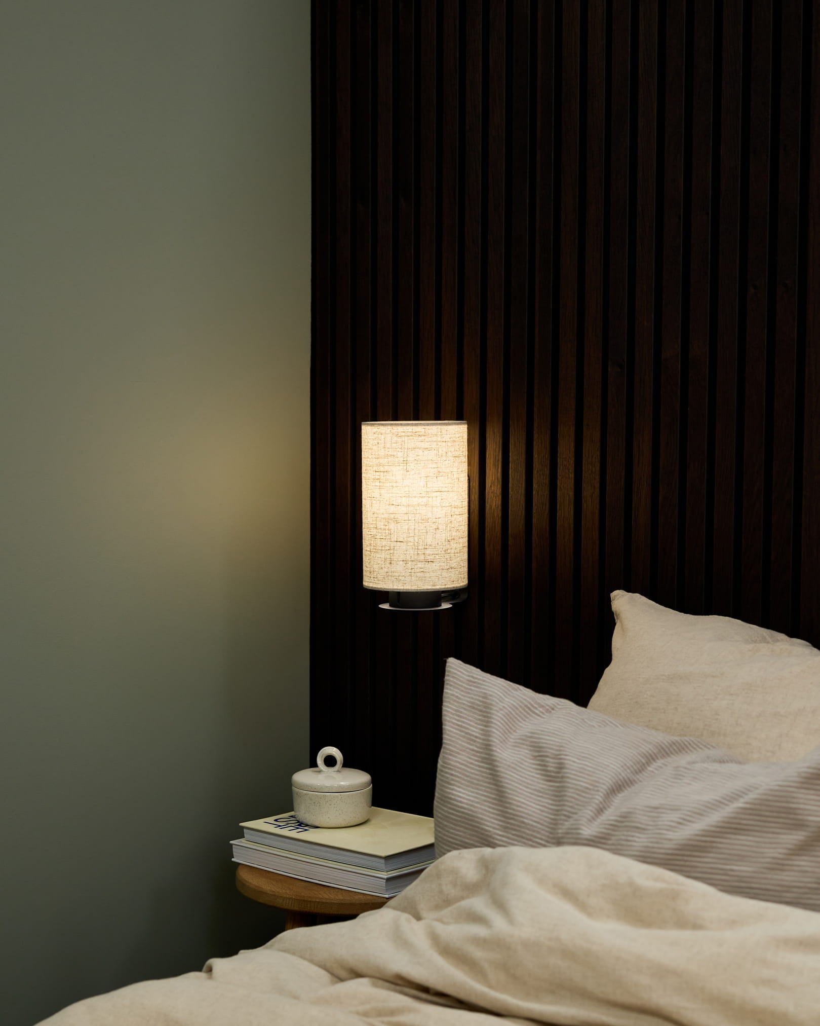 Slaapkamerhoek met een WoodUpp Feya wandlamp in Black/Beige, beddengoed in beige en een nachtkastje met boeken en een keramische beker met deksel.