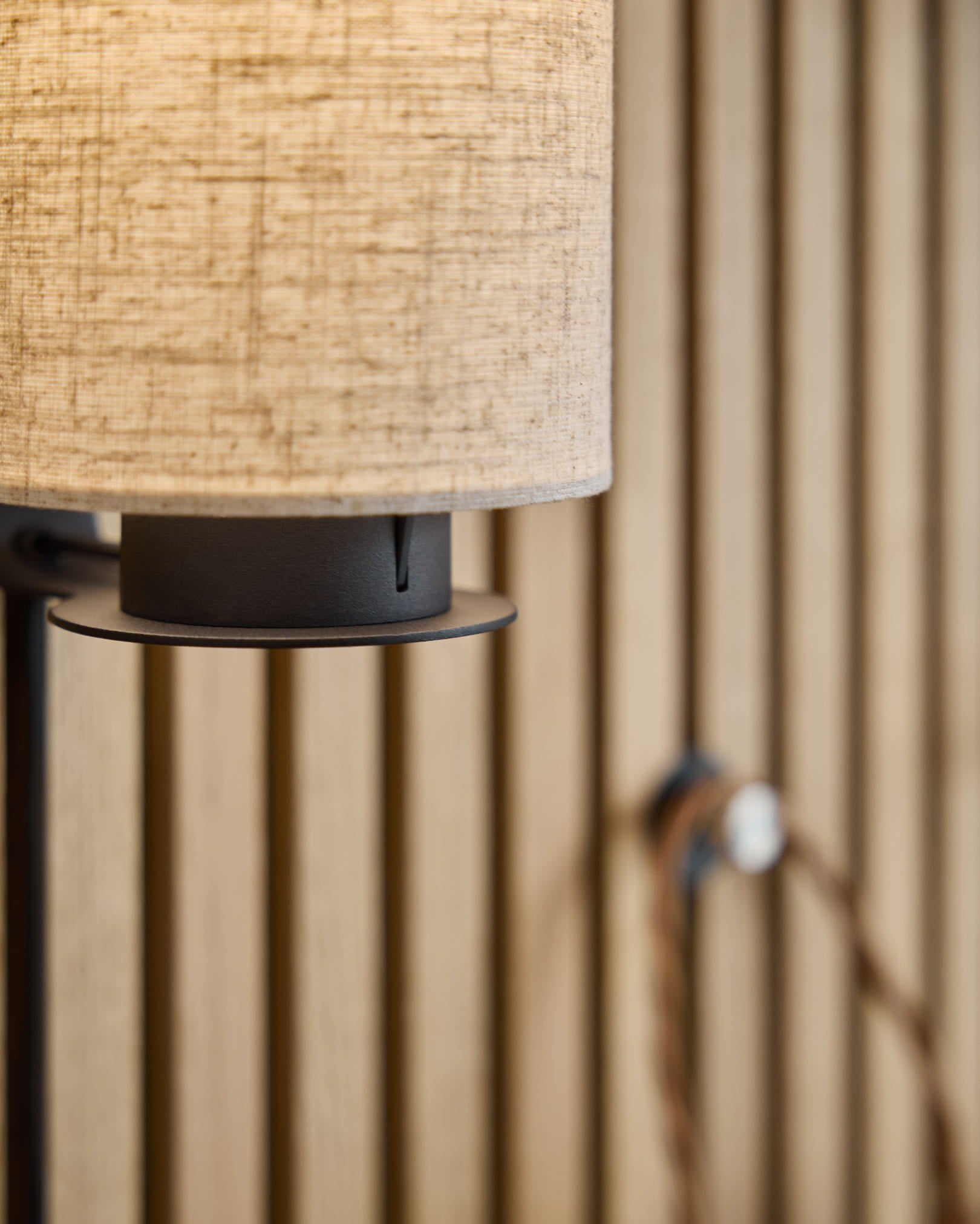 Feya wandlamp / Black/Beige van WoodUpp met een kap van linnen-katoen in beige, een metalen voet in black en een zichtbaar snoer, gemonteerd op een Scandinavisch akoestisch lattenwandpaneel.