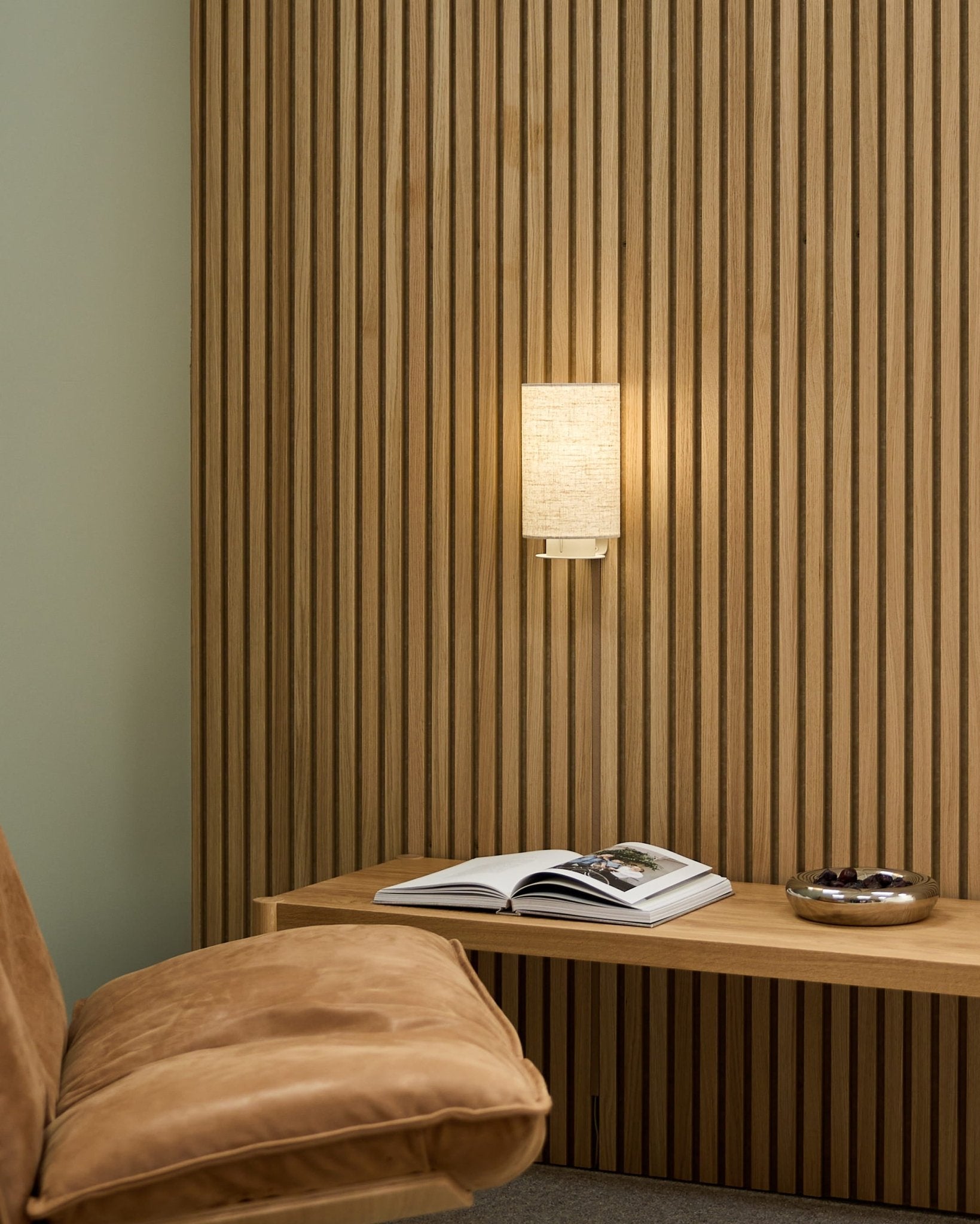 Pièce nordique minimaliste avec une chaise fauve, un mur acoustique à lattes WoodUpp Akupanel, l’applique Feya en Beige/Beige, un livre ouvert sur un banc et un bol.