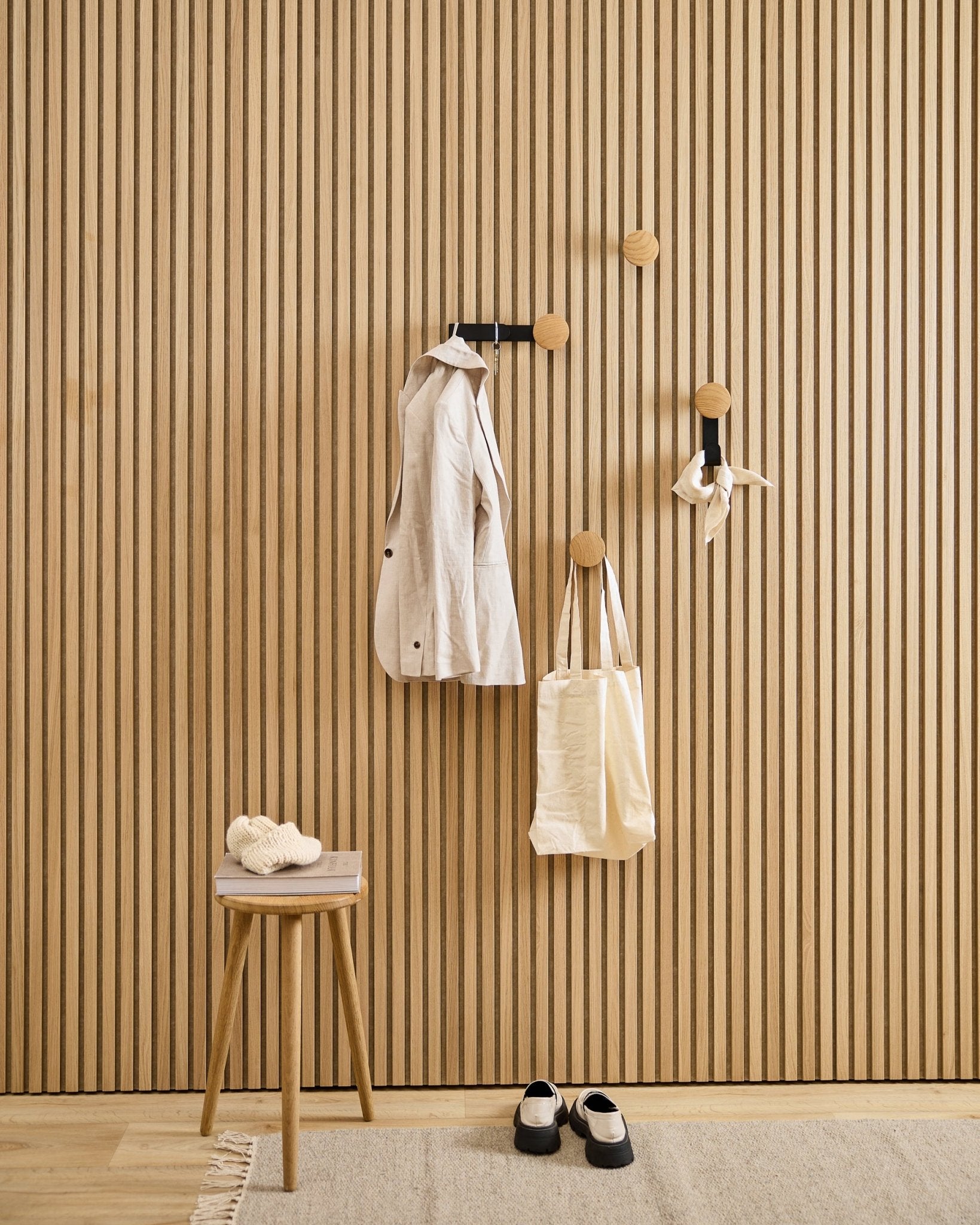 Minimalistische hal met de WoodUpp Linn Vertical Hanger in natural oak, met een jas van beige, een shopper, een sjaal, een kruk met pantoffels en schoenen in black en wit.