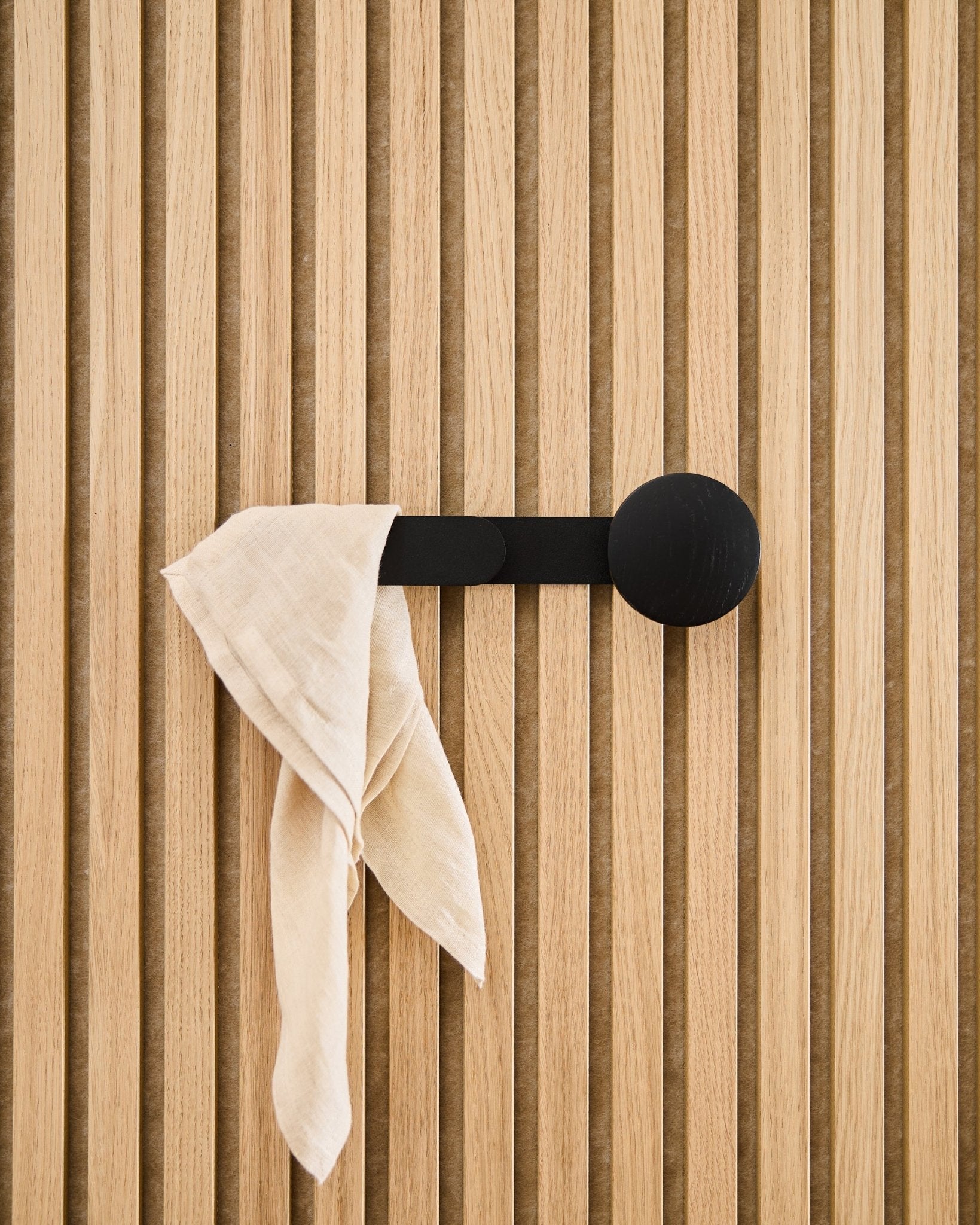 Een beige doek hangt over de WoodUpp Linn Horizontal Hanger in Black Oak, bevestigd aan een vertical akoestisch lattenwandpaneel.