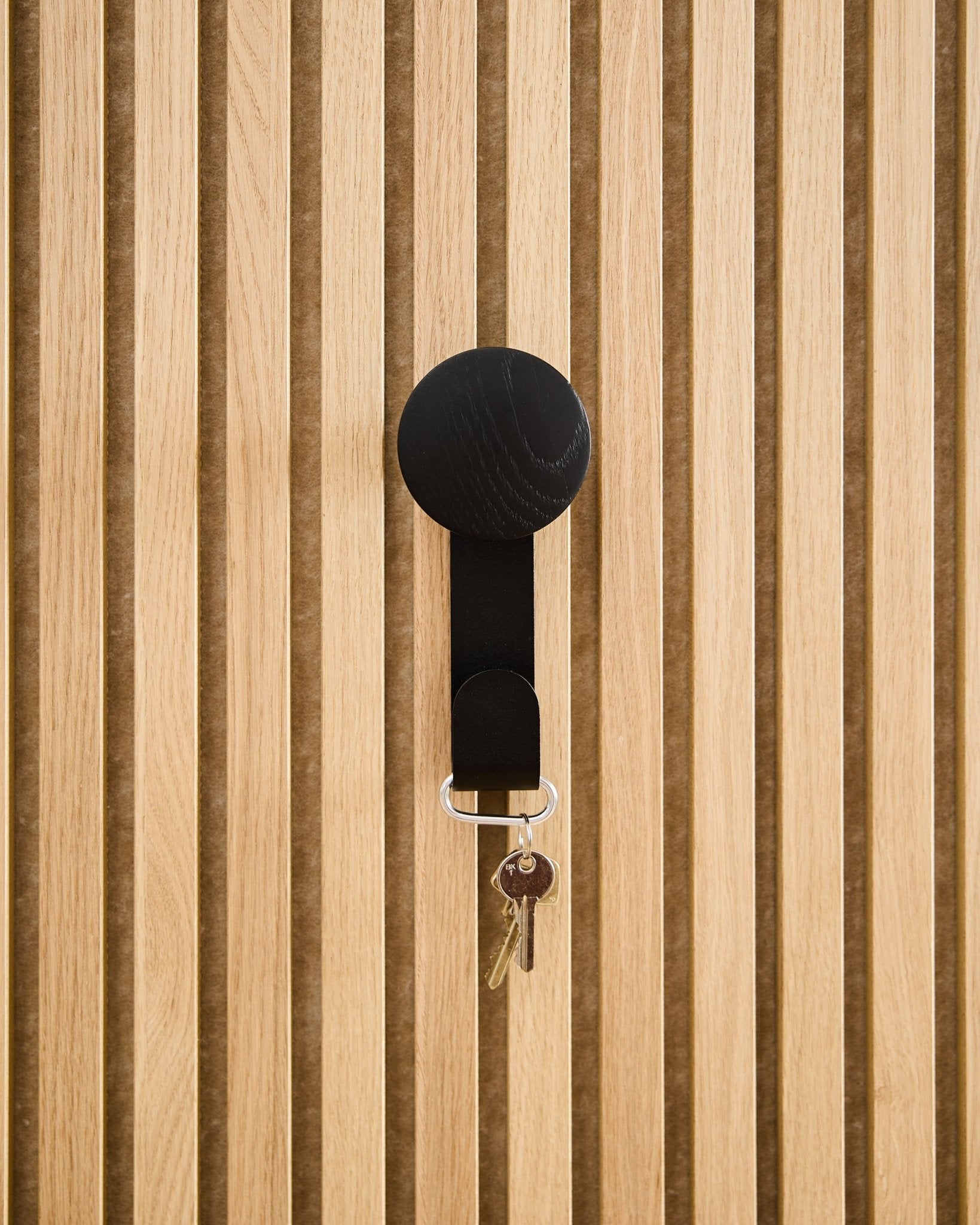 Een Linn Vertical Hanger in Black Oak van WoodUpp houdt een sleutelbos met twee sleutels en een ronde tag vast, gemonteerd op een paneel van Akupanel akoestisch hout.
