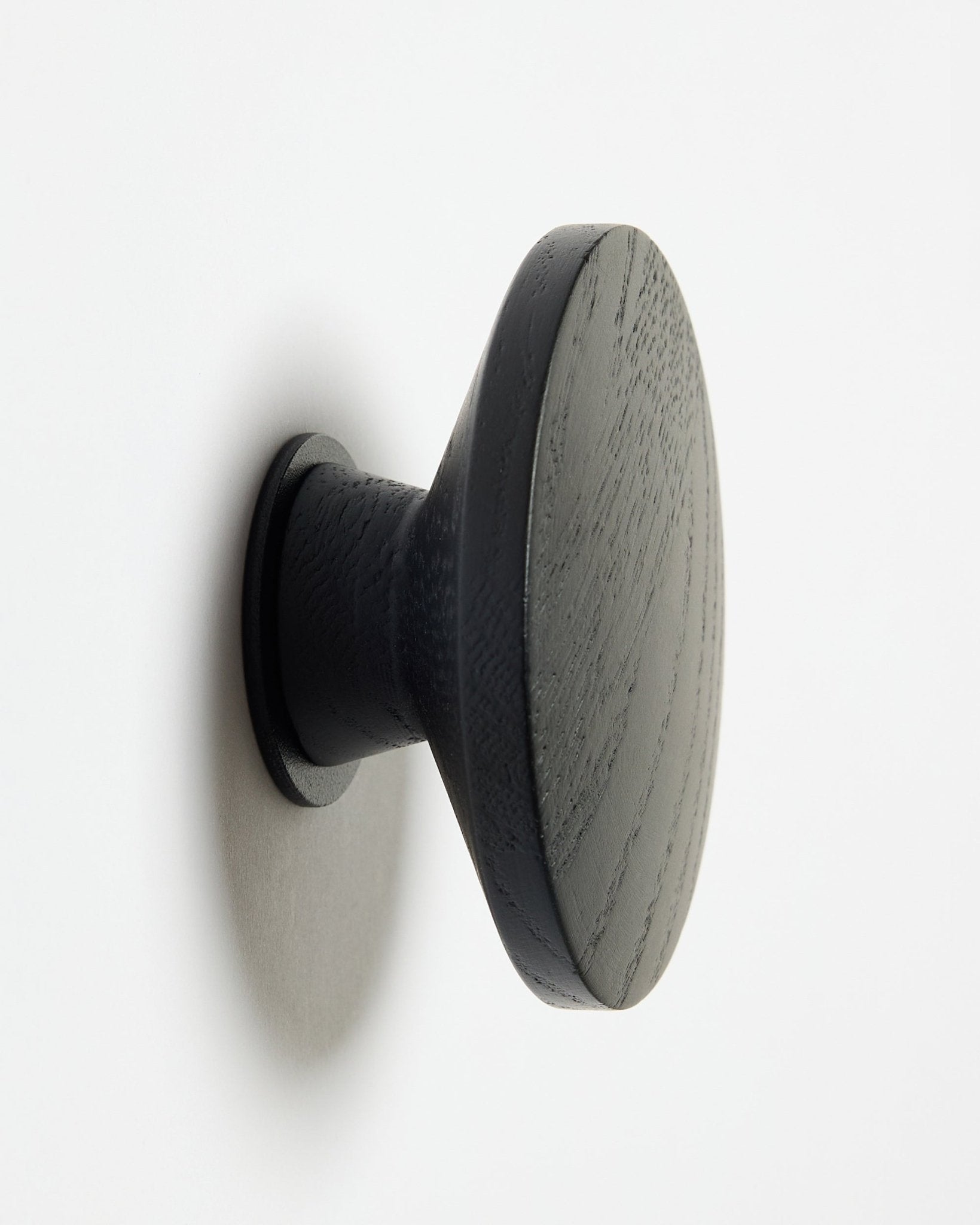 Een ronde, black oak Linn | Single Hanger van WoodUpp, ontworpen als accessoire voor Akupanel akoestische houten panelen, gemonteerd op een wit oppervlak met een zachte schaduw aan de linkerkant.