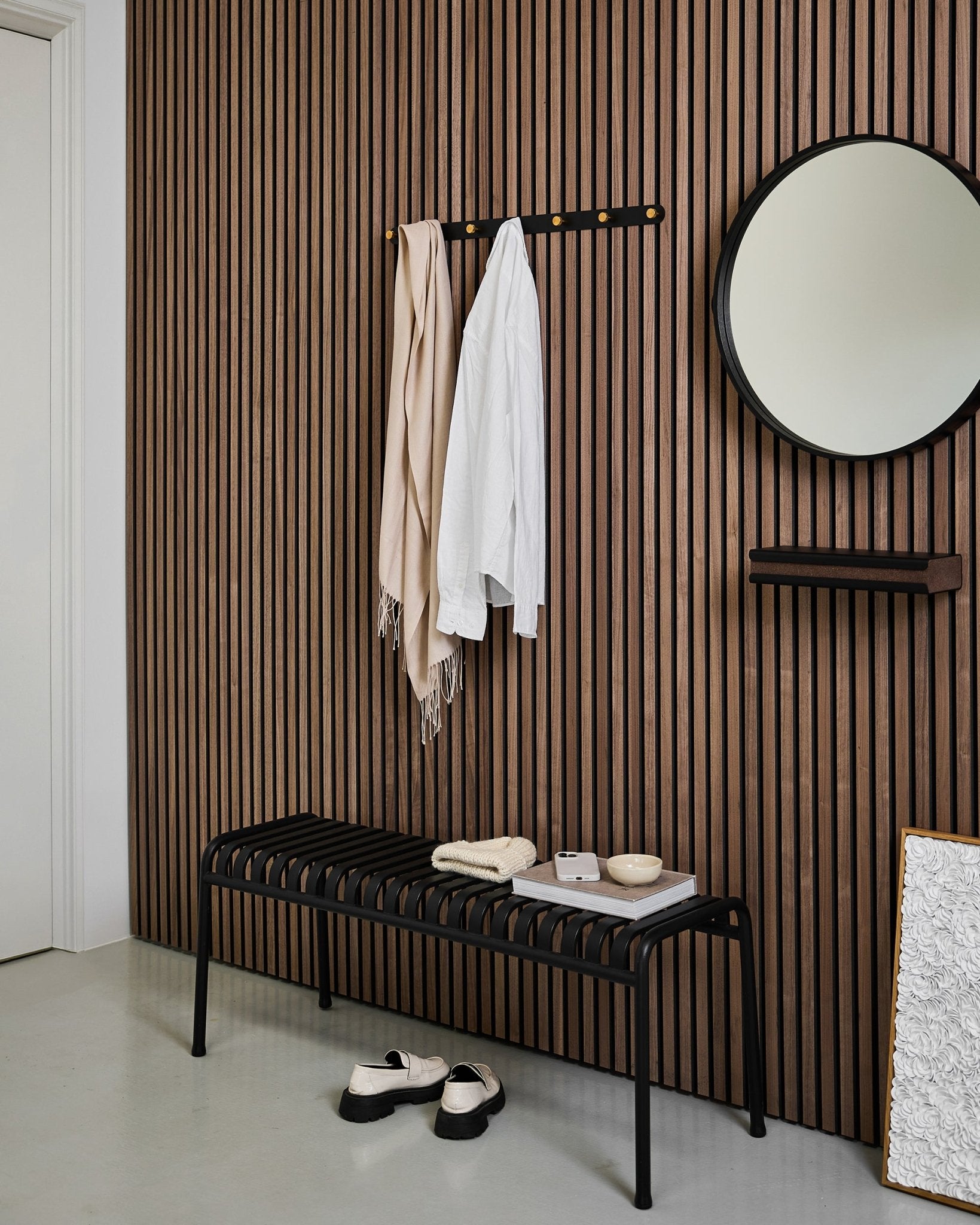 Minimalistische entree met een WoodUpp Akupanel akoestische lattenwand, Norr kapstok (Black/Brass, X7), bankje, spiegel en moderne decoratie in neutrale tinten.