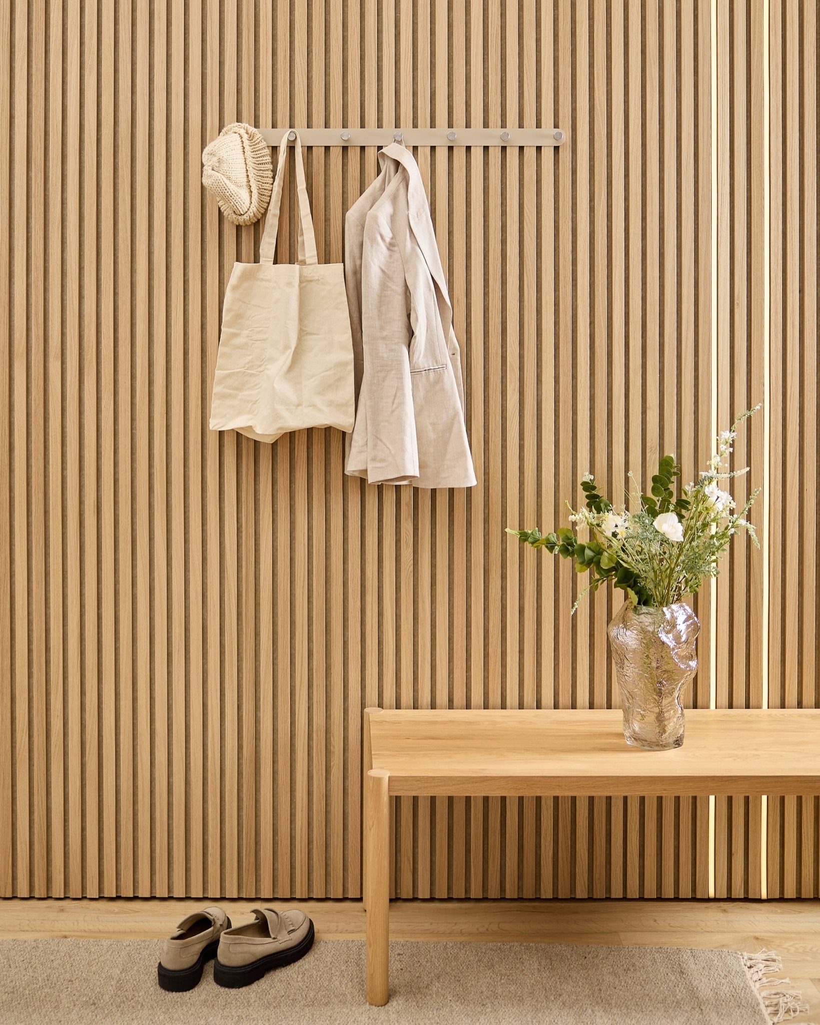 Een beige tas, hoed en shirt hangen aan de WoodUpp Norr kapstok boven schoenen, met daarnaast een bankje en vaas naast het gestreepte akoestische lattenwandpaneel.