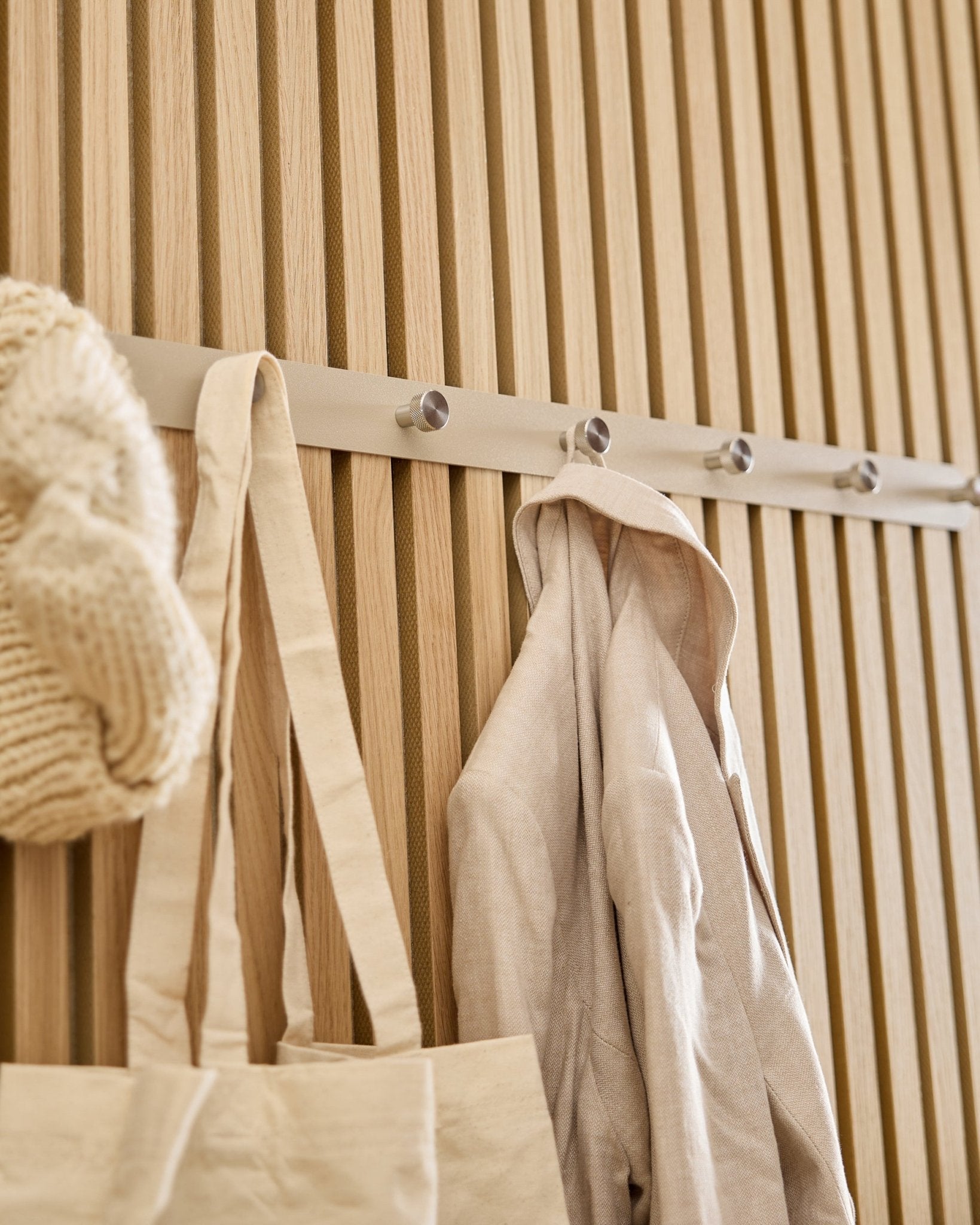 Een beige draagtas, gebreide muts en linnen shirt hangen aan de WoodUpp Norr kapstok (Beige/roestvrij Steel, X7), gemonteerd op een licht houten Akupanel lattenwand.