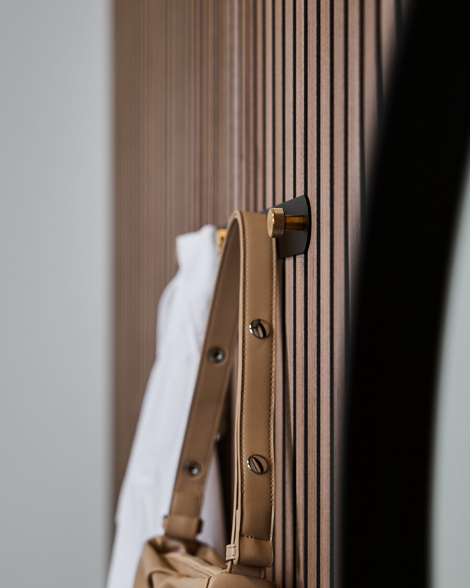 Een lichtbruine tas en wit overhemd hangen aan een WoodUpp Norr kapstok (Black/Brass, X5), gemonteerd op een vertical Akupanel akoestisch lattenwandpaneel.