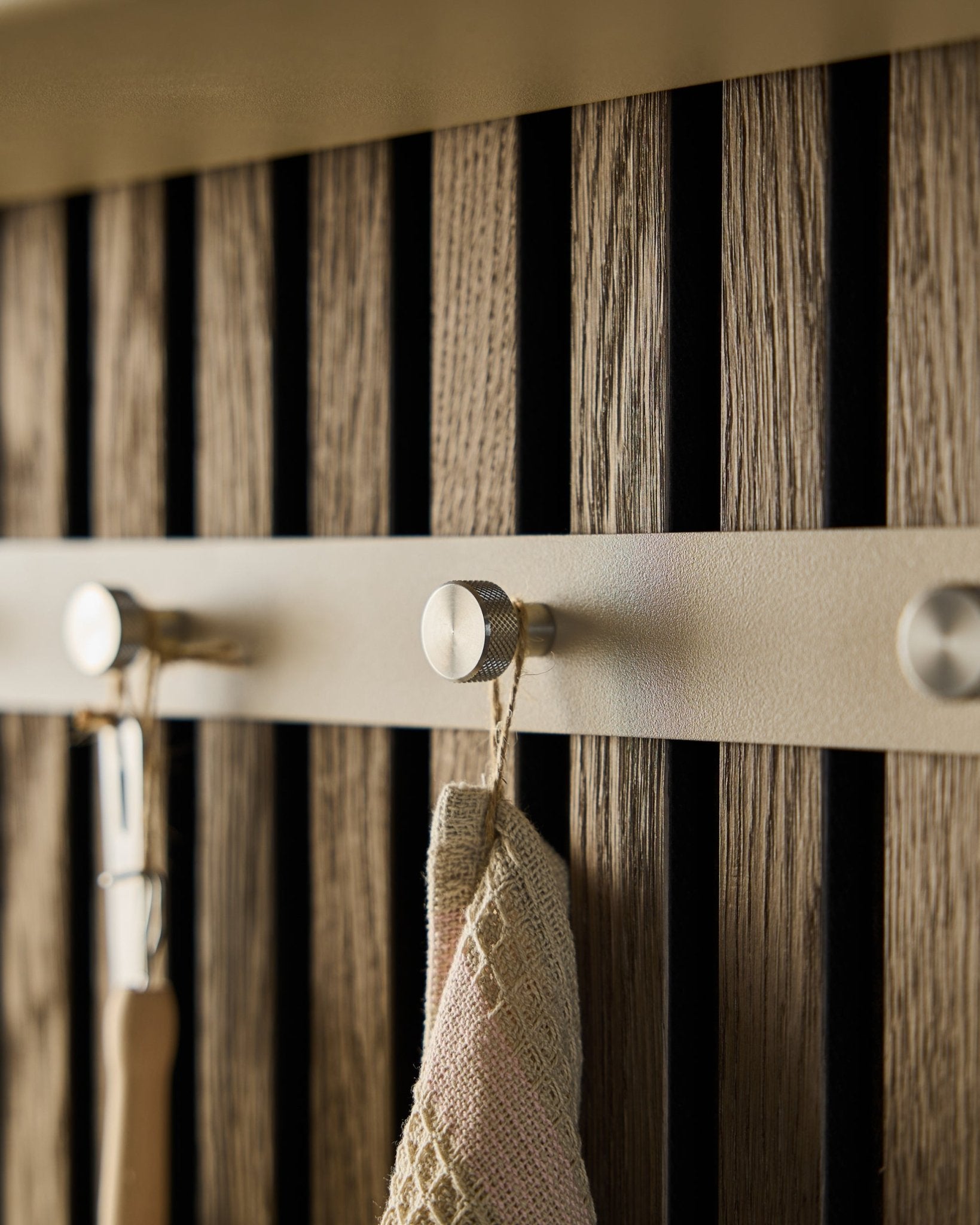 Gros plan sur le porte-manteau mural Norr de WoodUpp (Beige/acier inoxydable Steel, X5) monté sur un panneau à lattes en bois Panneau acoustique, avec une serviette et un outil à manche suspendus.