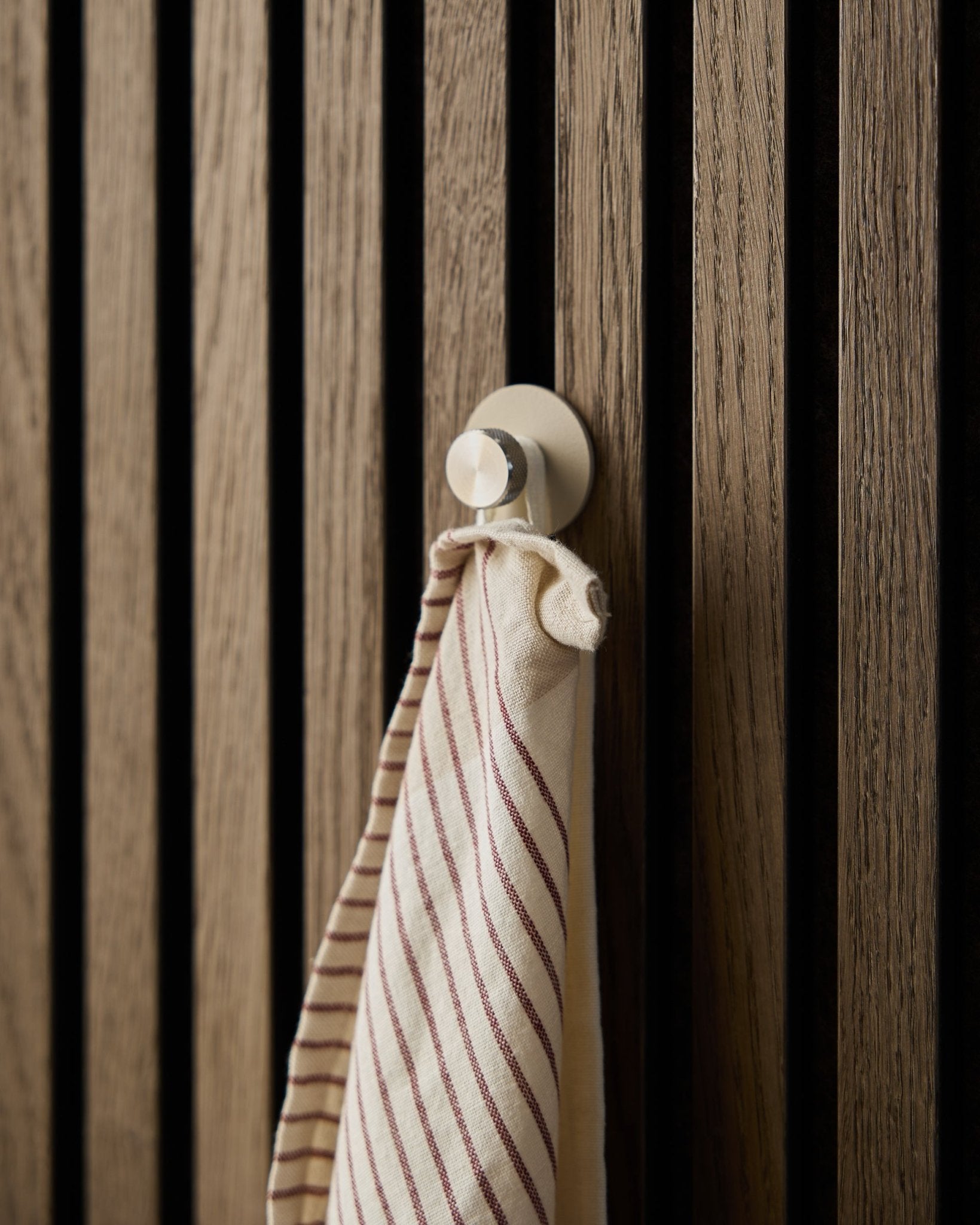 Een gestreepte handdoek hangt aan een beige Norr kapstokhaak van WoodUpp, gemaakt van roestvrij steel, gemonteerd op een akoestisch lattenwandpaneel van hout met textuur in Akupanel.