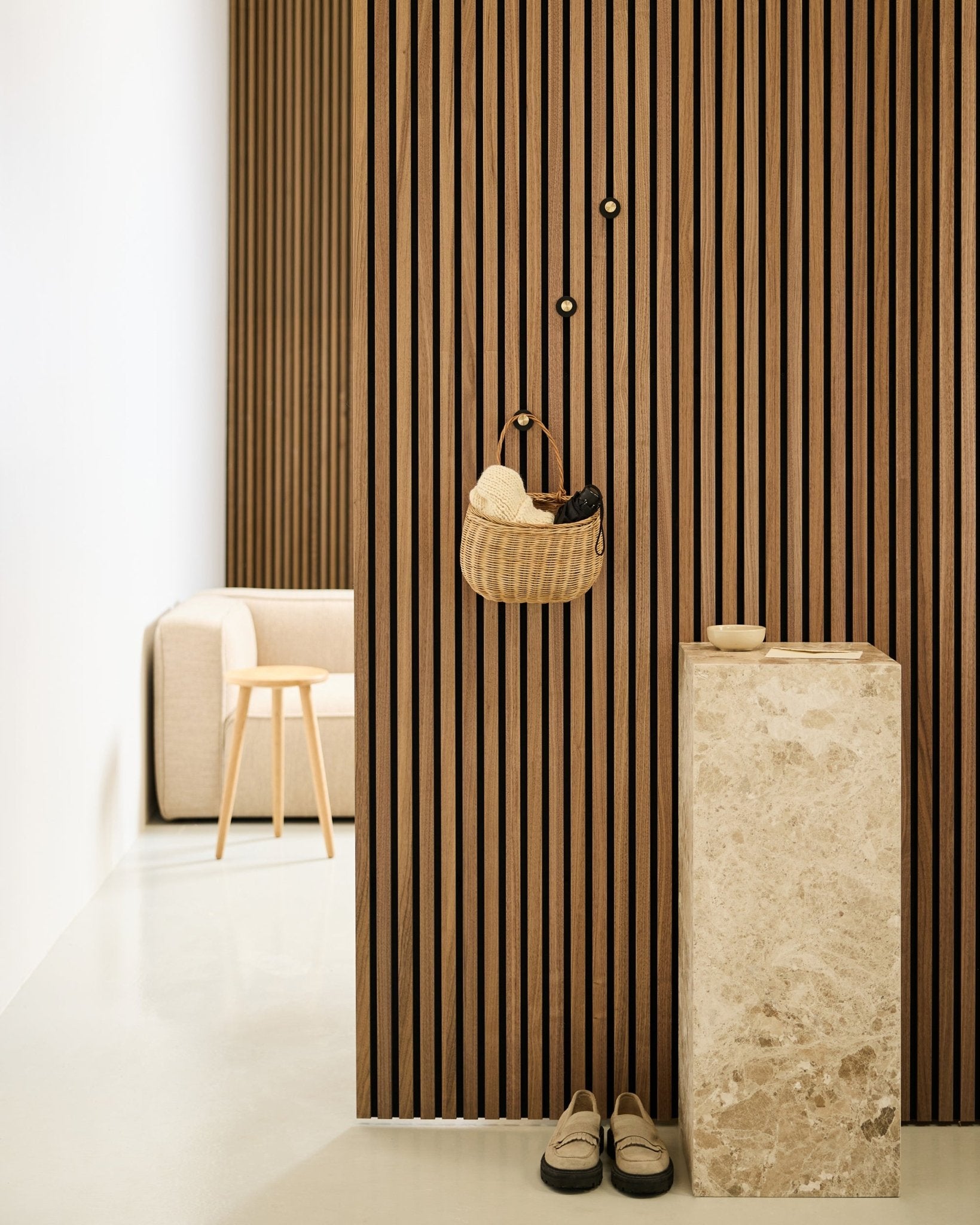 Minimalistische hal met Akupanel akoestische lattenwand, WoodUpp Norr kapstokhaak in black en brass, mand, stenen sokkel, pantoffels en beige stoel.