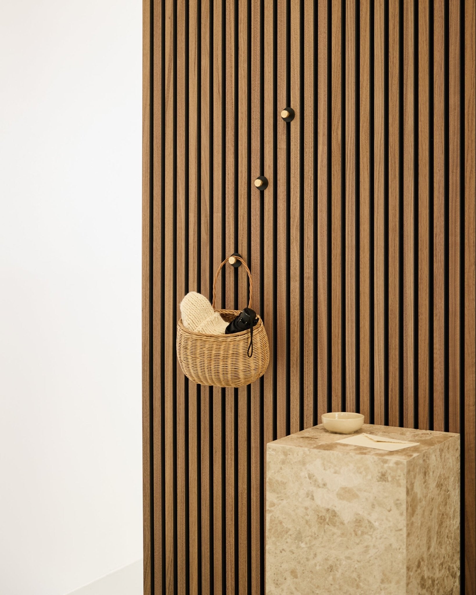 Patère Norr de WoodUpp montée sur un mur à lattes acoustiques nordique Akupanel, à côté d’un piédestal en pierre beige avec un bol small dans un intérieur minimaliste.