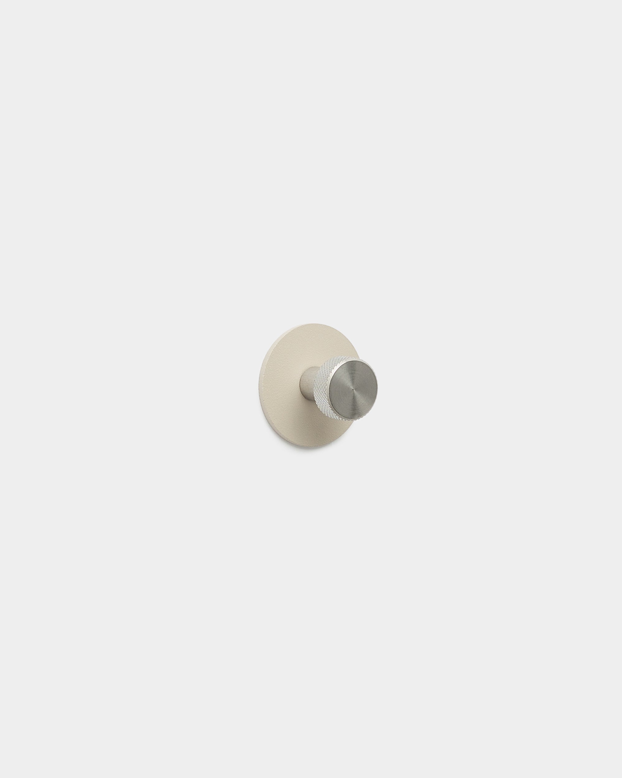 De WoodUpp Norr kapstokhaak in beige en roestvrij steel heeft een minimalistische haak en ronde basis, is gemaakt van gepoedercoat steel en maakt deel uit van de accessoires van WoodUpp.